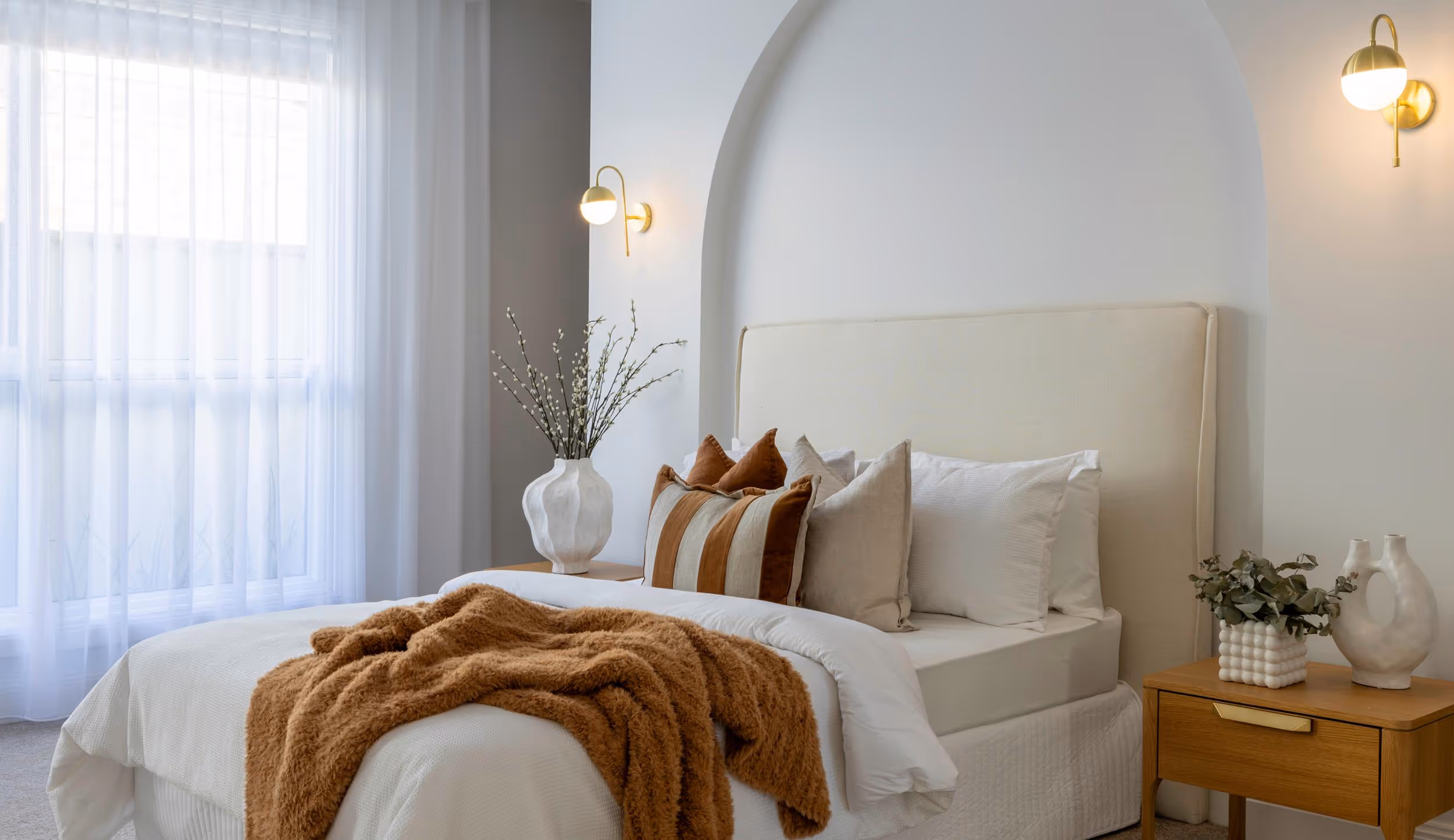 Modern bedroom with beige upholstered bed, white bedding, brown throw blanket, decorative pillows, wooden nightstands, and soft wall sconces.
