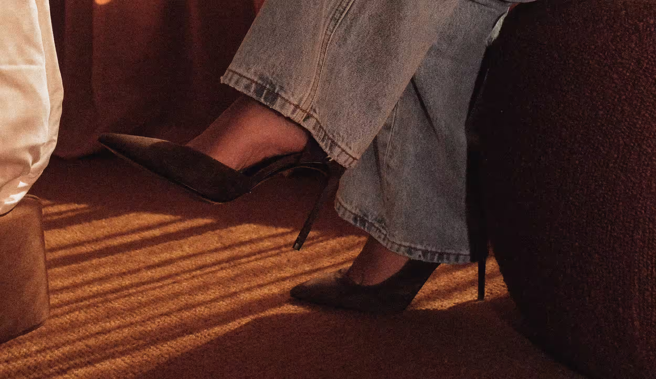 Person wearing light blue jeans and black high-heeled shoes sitting on a brown couch with sunlight casting shadows on a brown carpet.
