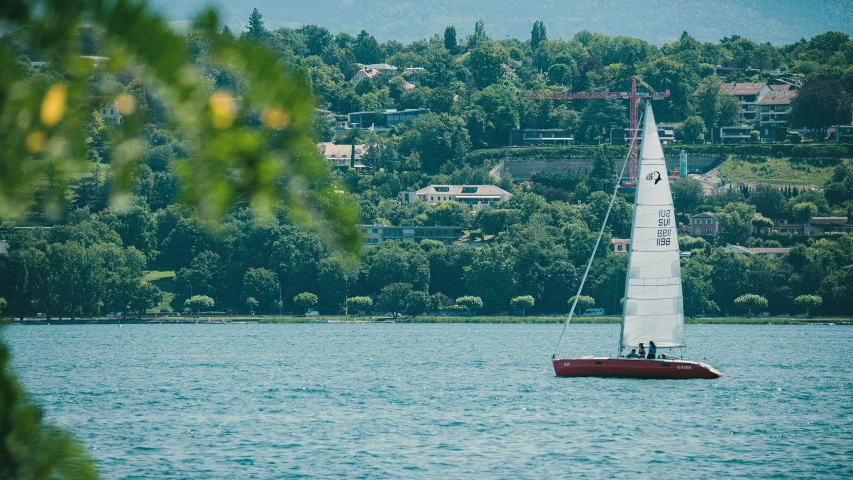 Voilier blanc avec des personnes naviguant sur un lac bordé d'une forêt et avec des maisons en arrière-plan.