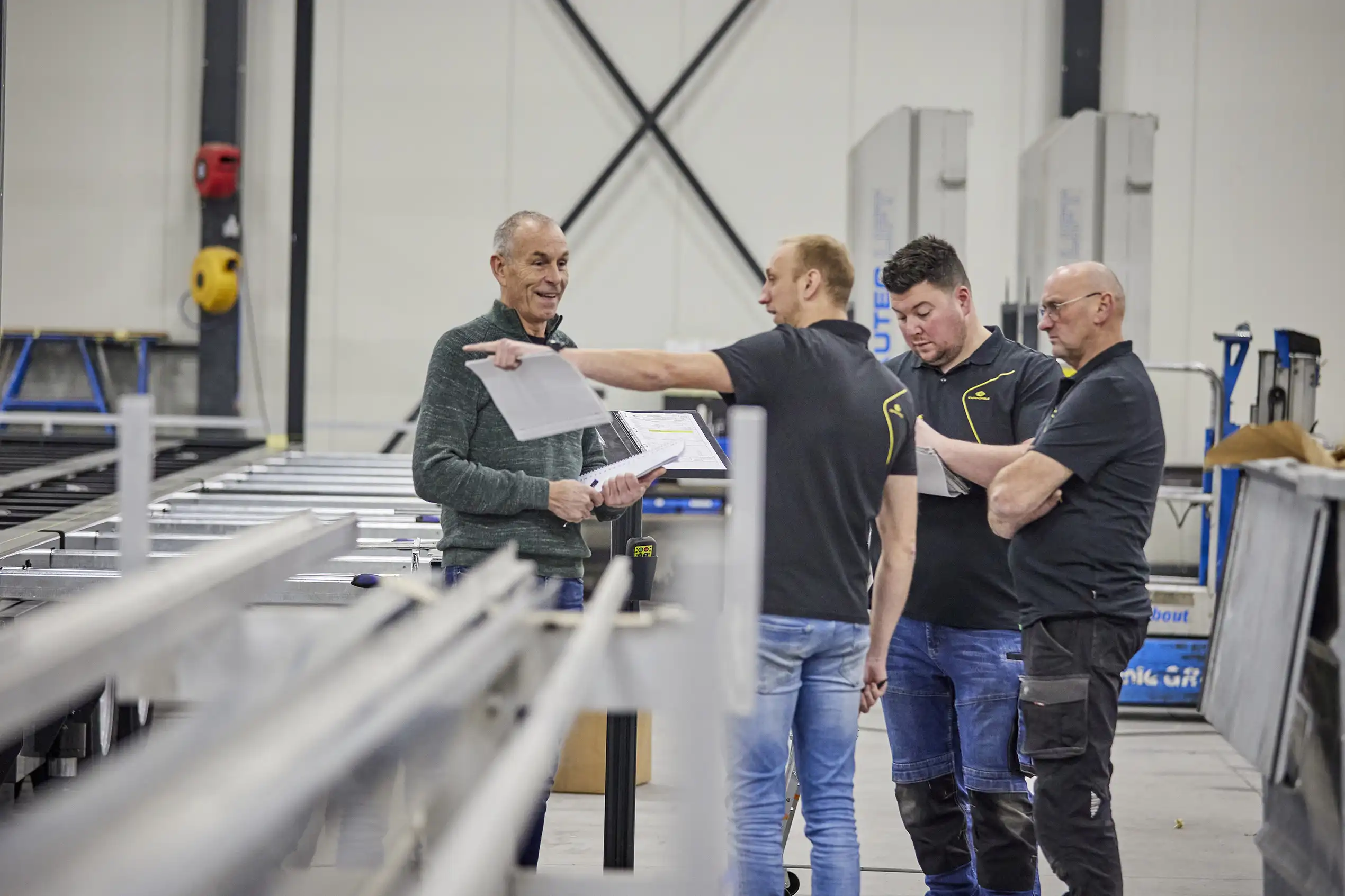 Team members in a high-tech manufacturing facility discussing over a blueprint. The senior member points something out on the paper to others in a collaborative environment. The background shows the intricate assembly line for expandable mobile spaces.