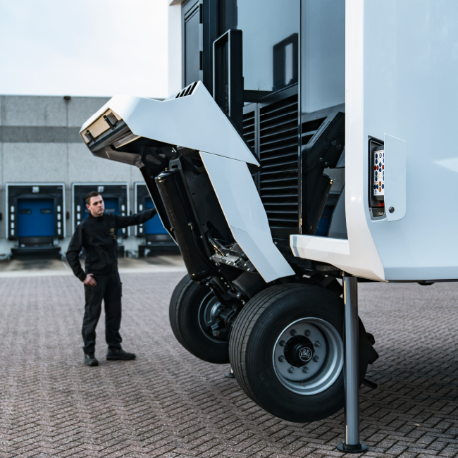 A man in work attire stands next to a large, innovative mobile structure with its front section lifted, revealing complex mechanisms underneath.