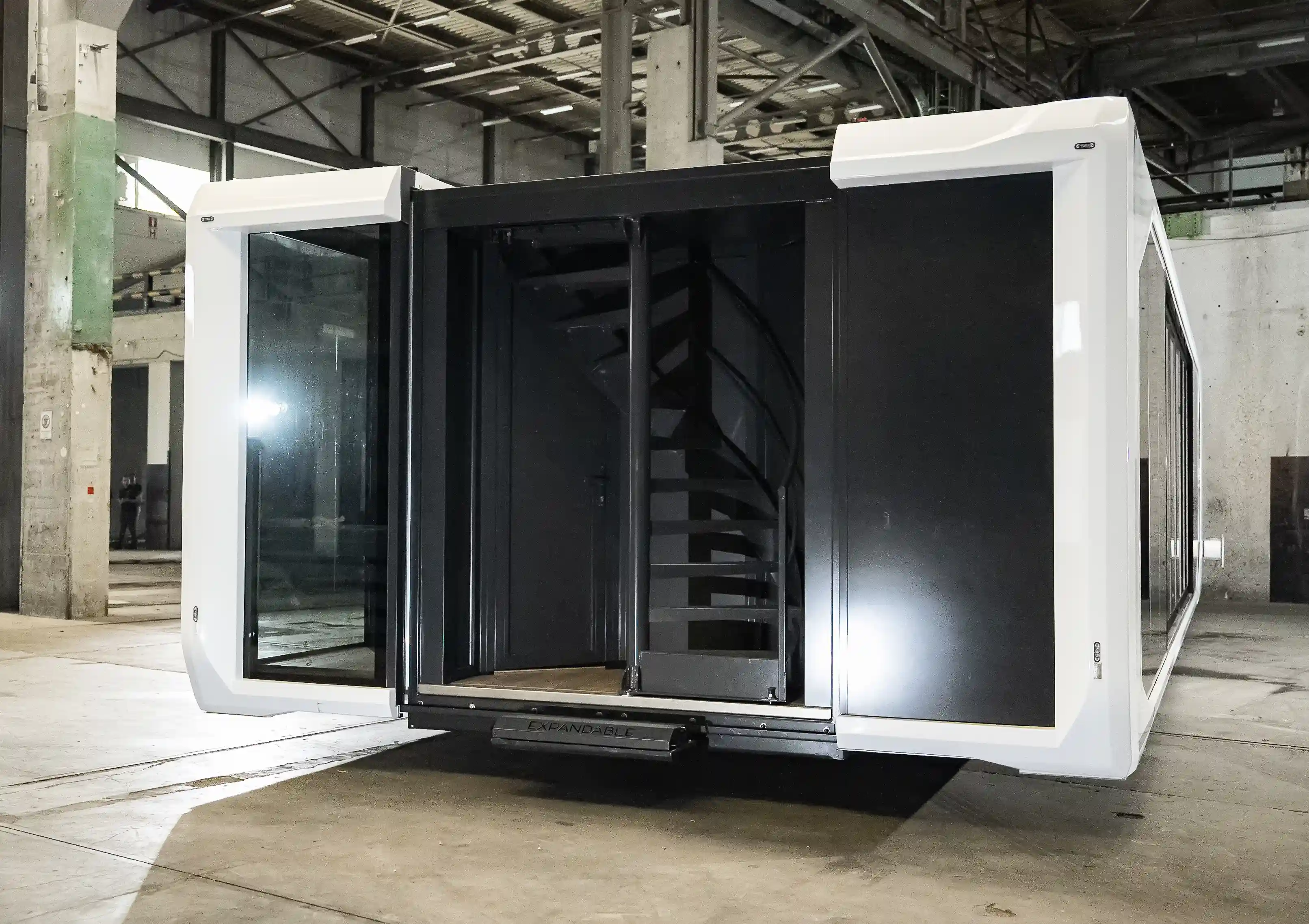 Open entry of an Expandable relocatable space unit featuring a modern spiral staircase for rooftop access, shown inside an industrial hall