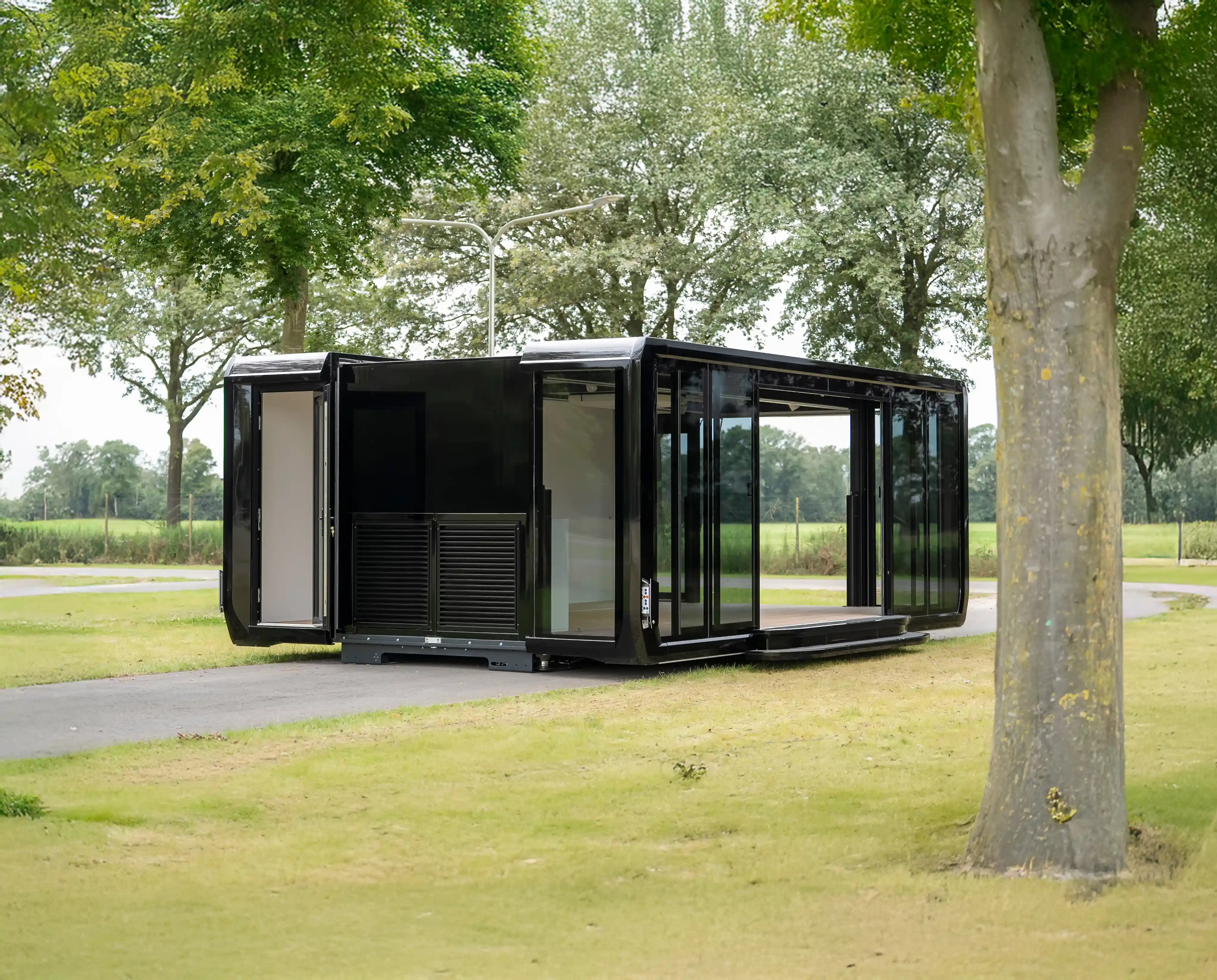 Modern black expandable pod set up on a paved surface in a grassy, tree-lined outdoor area, with both sides extended to showcase its spacious interior and glass sliding doors.