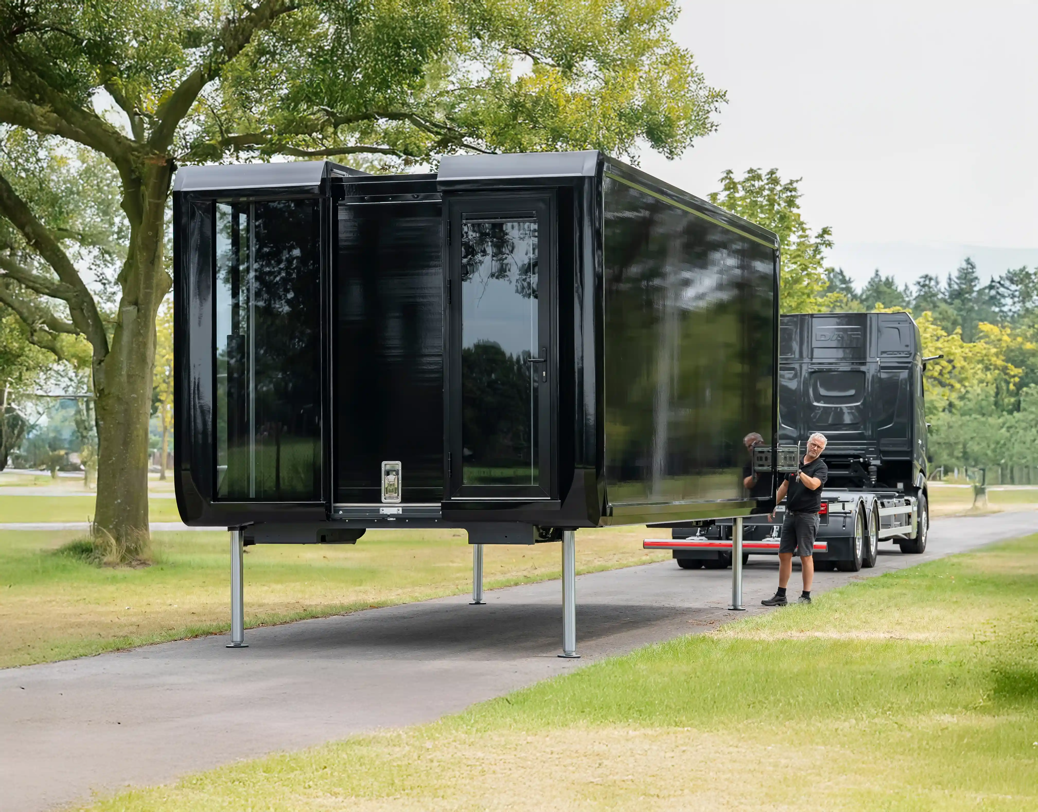 Expandable black pod being unloaded and set up on extendable metal legs by a worker next to a black transport truck, on a paved path surrounded by grassy fields and trees.