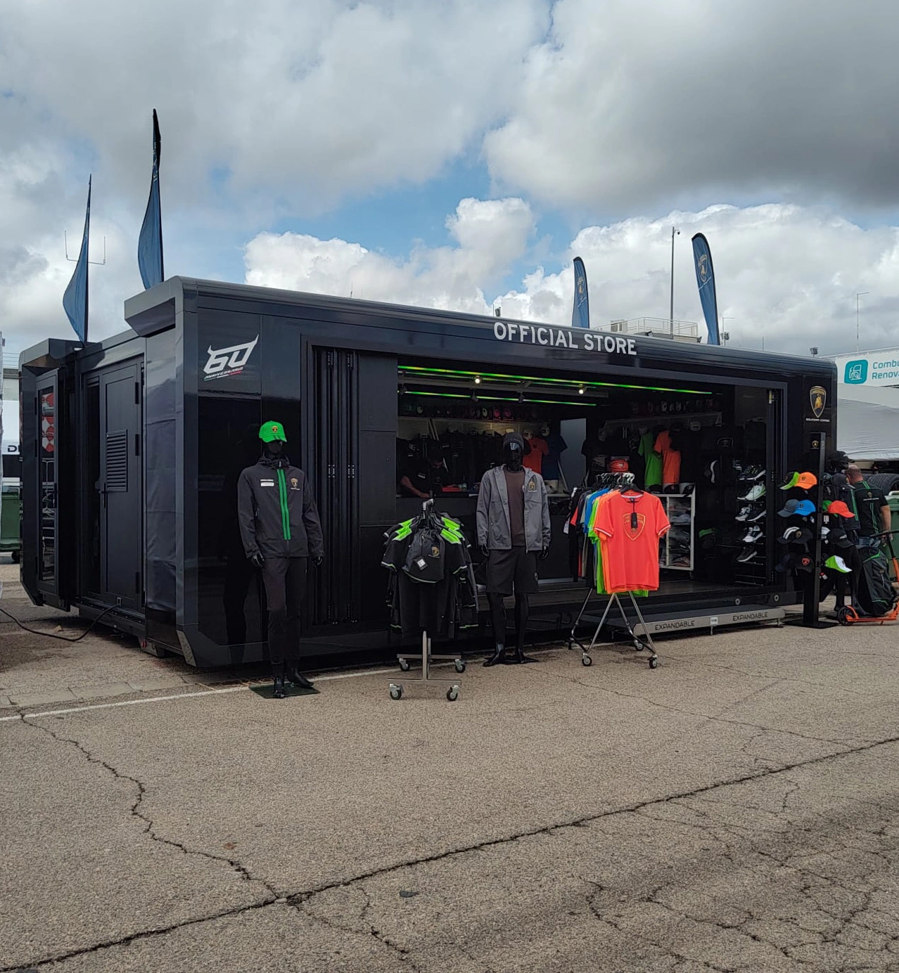 A black sports merchandise trailer with customers browsing clothing displayed outside under a cloudy sky.