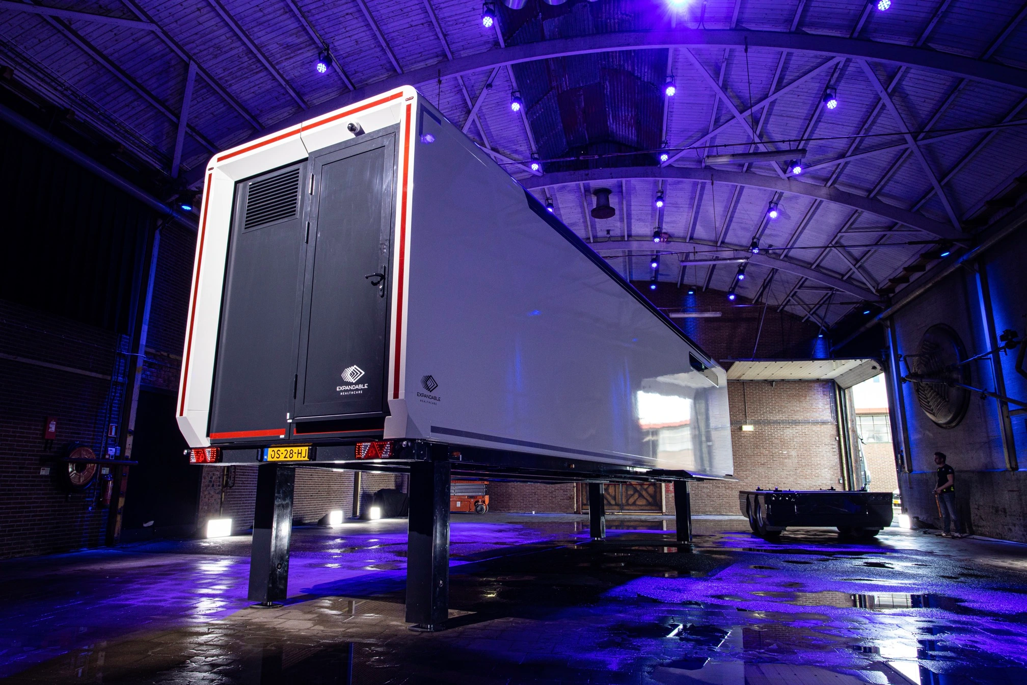 An emergency response trailer parked inside an industrial brick warehouse, lit by overhead blue lights with a person kneeling nearby handling equipment.