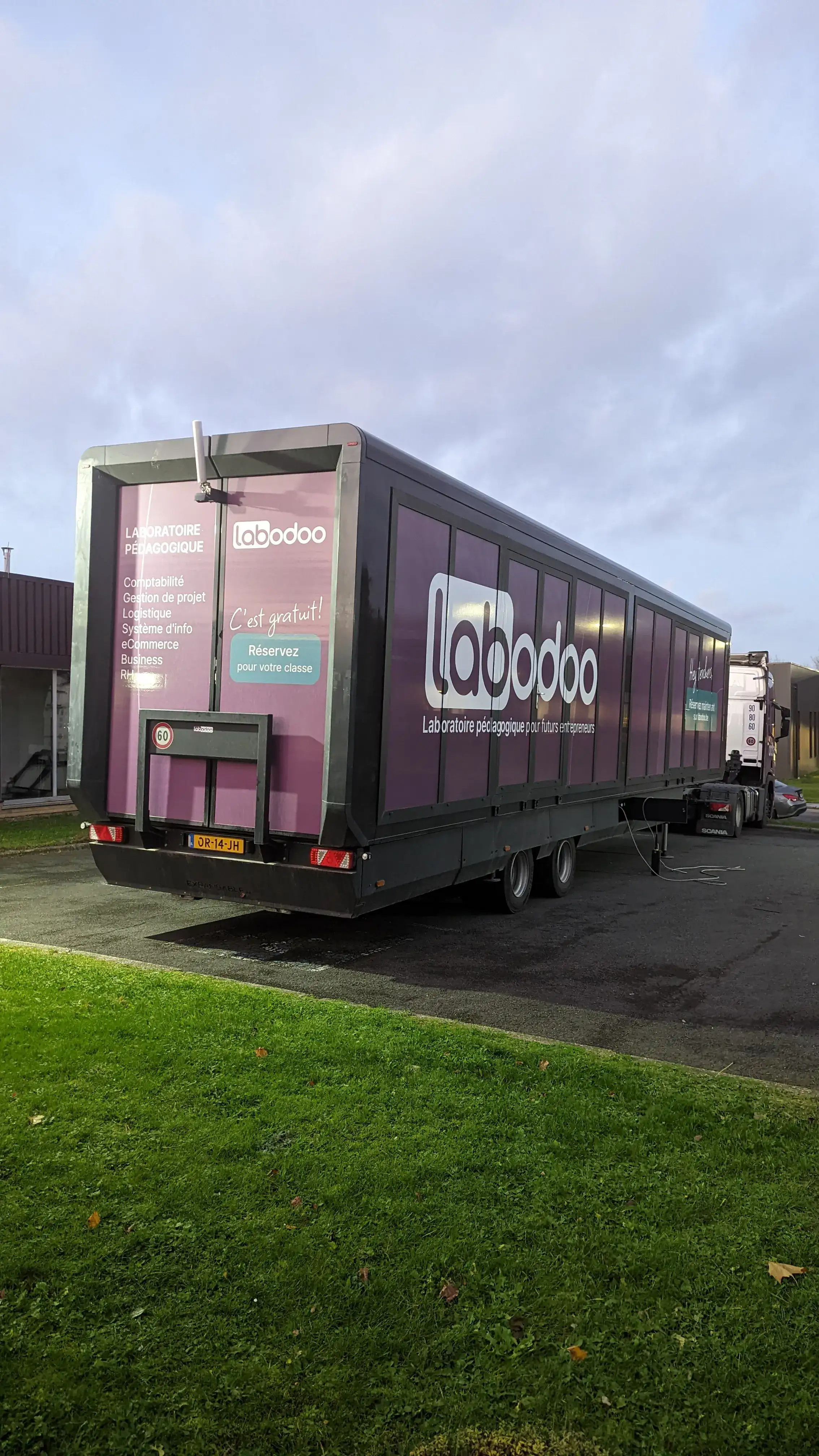 Une roulotte de classe mobile pour « labodoo » avec des escaliers déployés, prête à accueillir les élèves, sur fond de ciel nuageux et d'herbe verte en toile de fond.