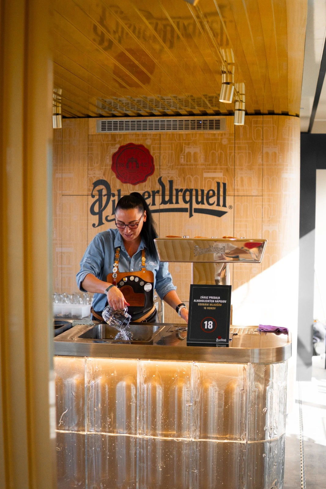 The inside of a promotional trailer with Pilsner Urquell branding, where a staff member is cleaning glasses inside a well-designed bar area.