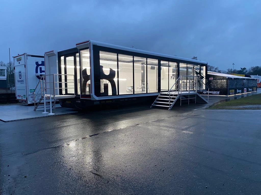 Escena crepuscular de un espacioso remolque de motocross con paredes transparentes, que muestra el interior y el logotipo de Husqvarna, en un lugar tranquilo para eventos.