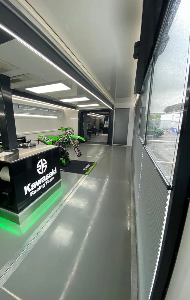 Vista interior de un remolque de motocross con un diseño elegante y moderno, con una bicicleta de carreras Kawasaki y la marca del equipo iluminada por luces de techo.