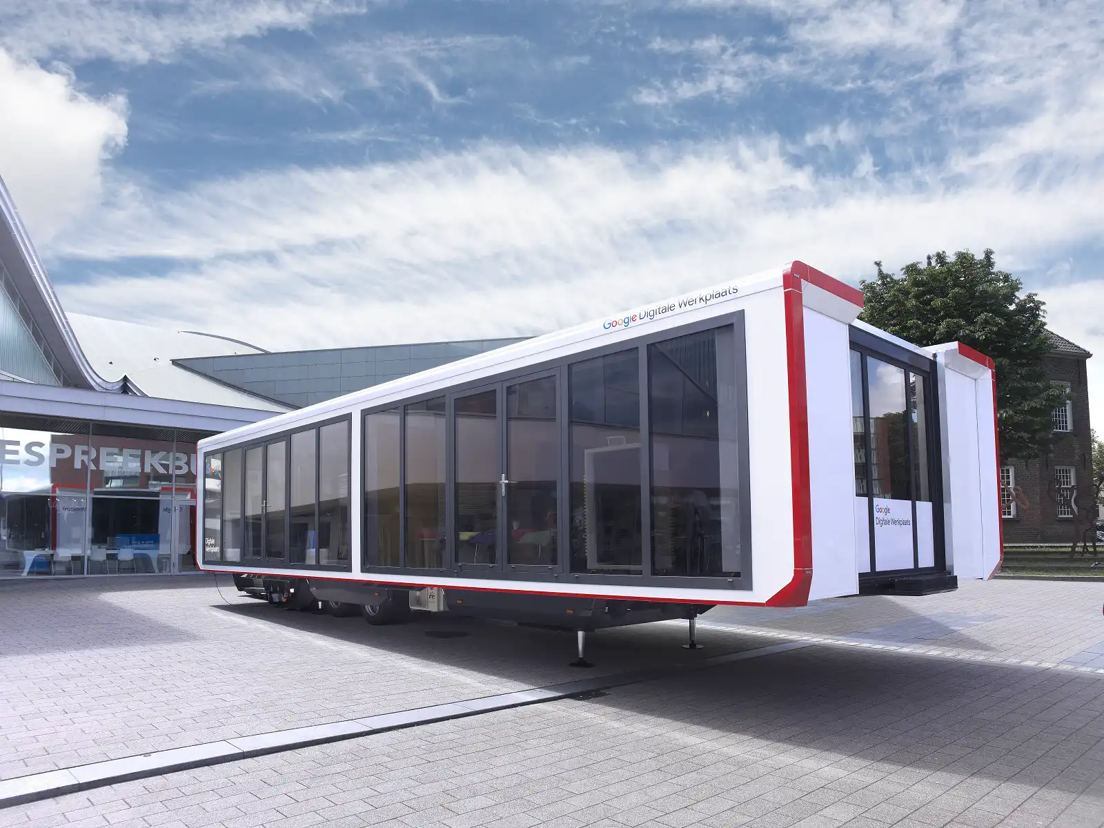 A modern advertising trailer with Google Digital Workshop branding, featuring glass walls and a clean, white and red design, parked in a plaza under a blue sky.