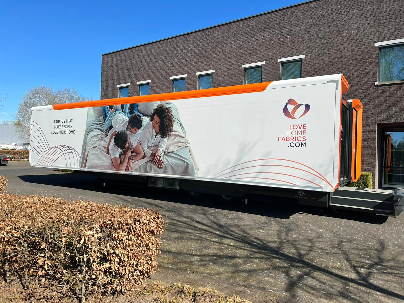 The exterior of a mobile showroom with 'LOVE HOME FABRICS' branding, designed with a clean, white body accented with orange and showcasing lifestyle imagery.