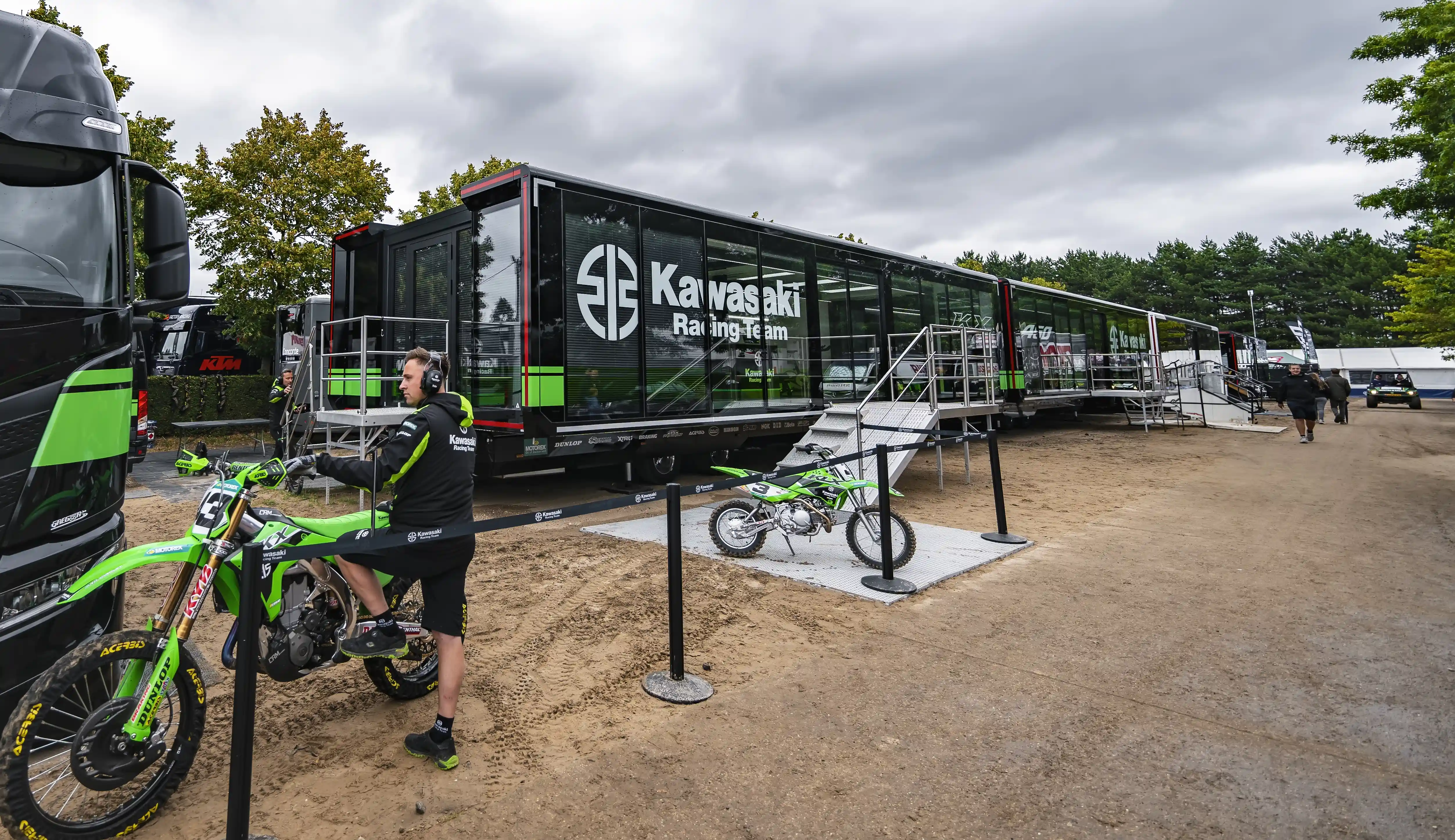 A view of a sports trailer for the Kawasaki Racing Team at a motocross event, with a green bike in front and the team's branding visible on the glass exterior.