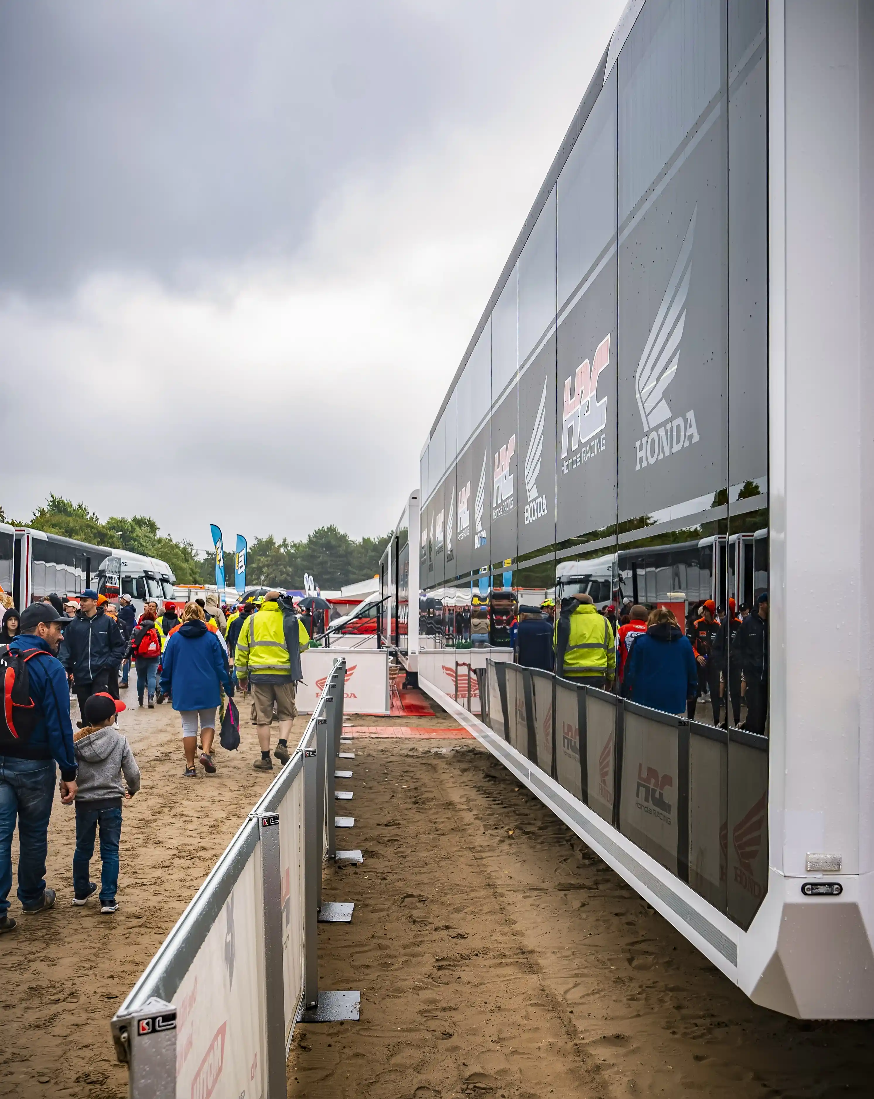 A sports trailer at an outdoor event, showcasing the HRC Honda team branding along its extended side, with visitors walking by on a sandy path.