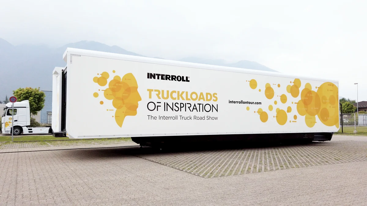 A large white semi-trailer parked in an open area with mountains in the background. The trailer has a bold design with the words 'INTERROLL TRUCKLOADS OF INSPIRATION' and 'The Interroll Truck Road Show' alongside a graphic of yellow-orange abstract shapes and splatters. The website 'interrollontour.com' is also displayed on the side of the trailer.