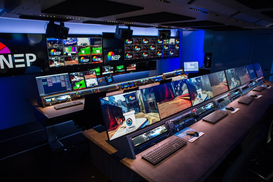 An advanced control room within an NEP broadcast trailer featuring a long desk with multiple monitors and broadcasting equipment, showcasing live feeds and operational software in a dimly lit environment.