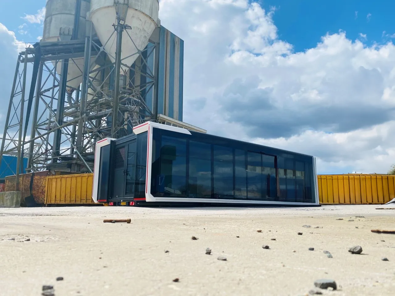 Expandable mobile hospitality trailer set against an industrial backdrop with a concrete silo, the trailer features a modern design with large windows and a bold red and white color scheme.