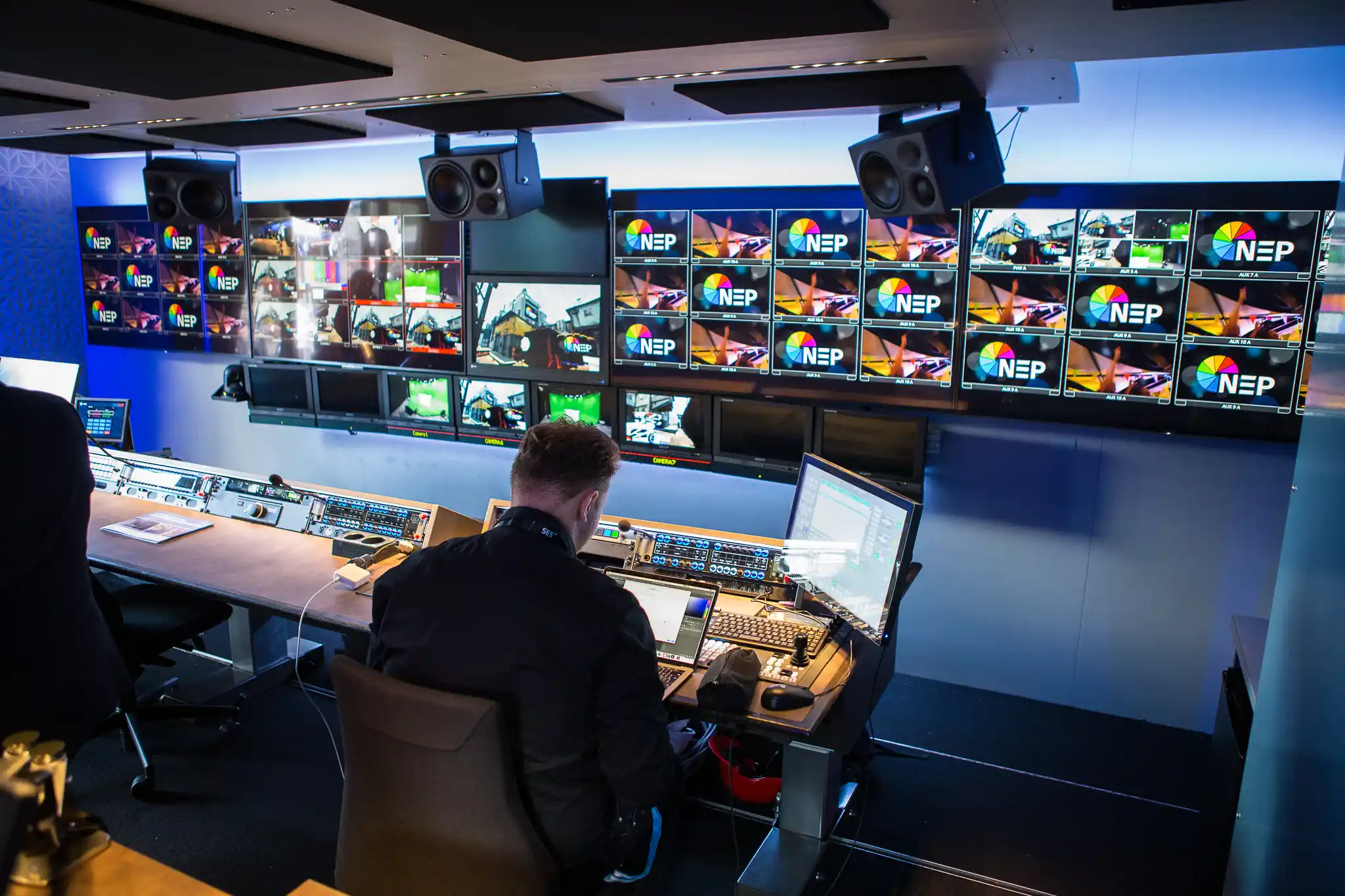 Inside view of a mobile control room with a person operating audio-visual equipment, surrounded by multiple screens displaying various camera feeds and graphics