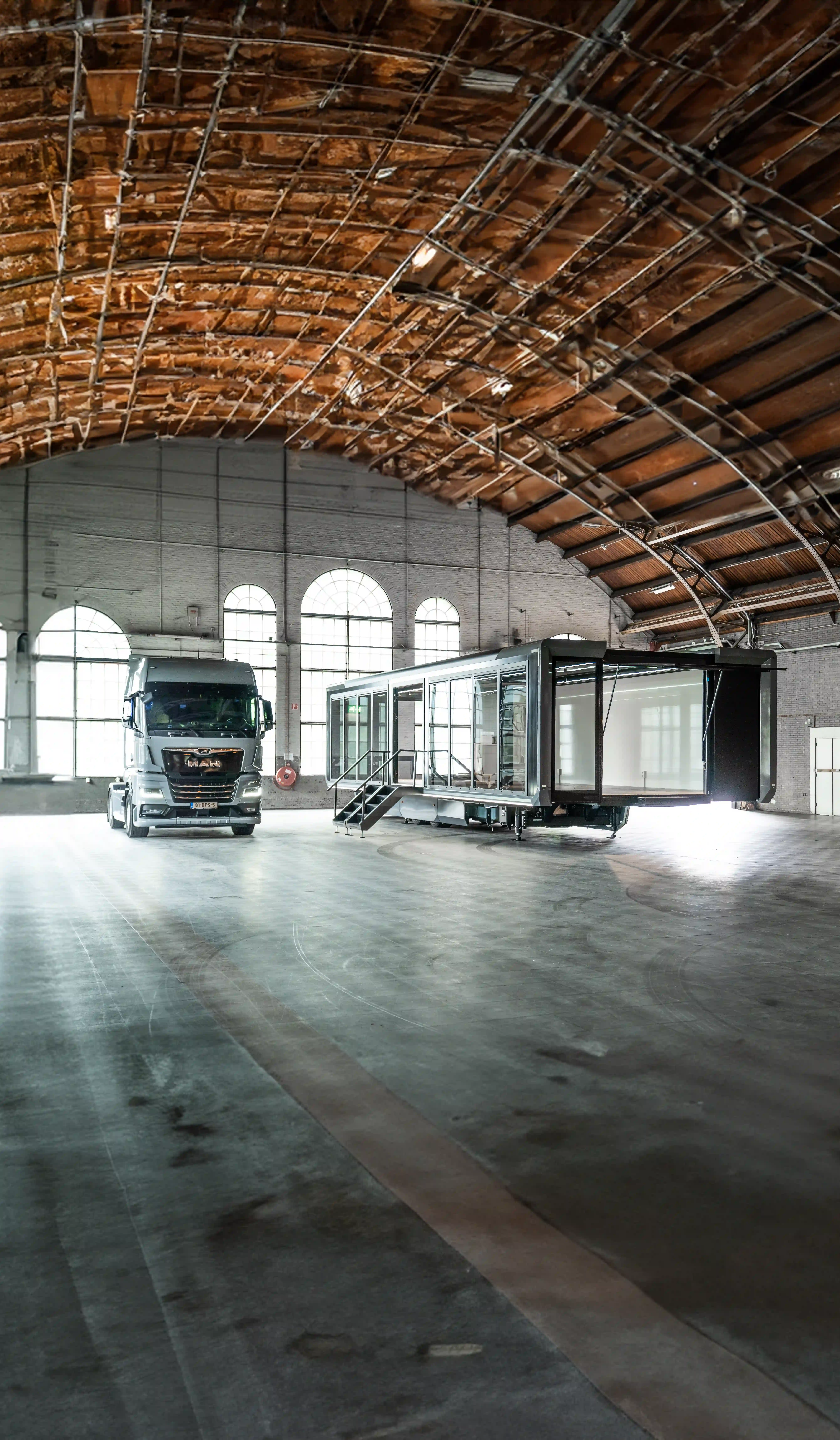 In einer weitlĂ€ufigen Industriehalle befindet sich ein mobiler Fotostudio-AnhĂ€nger neben einem groĂen LKW, was die PortabilitĂ€t des Studios und die einfache Einrichtung fĂŒr Aufnahmen vor Ort veranschaulicht.