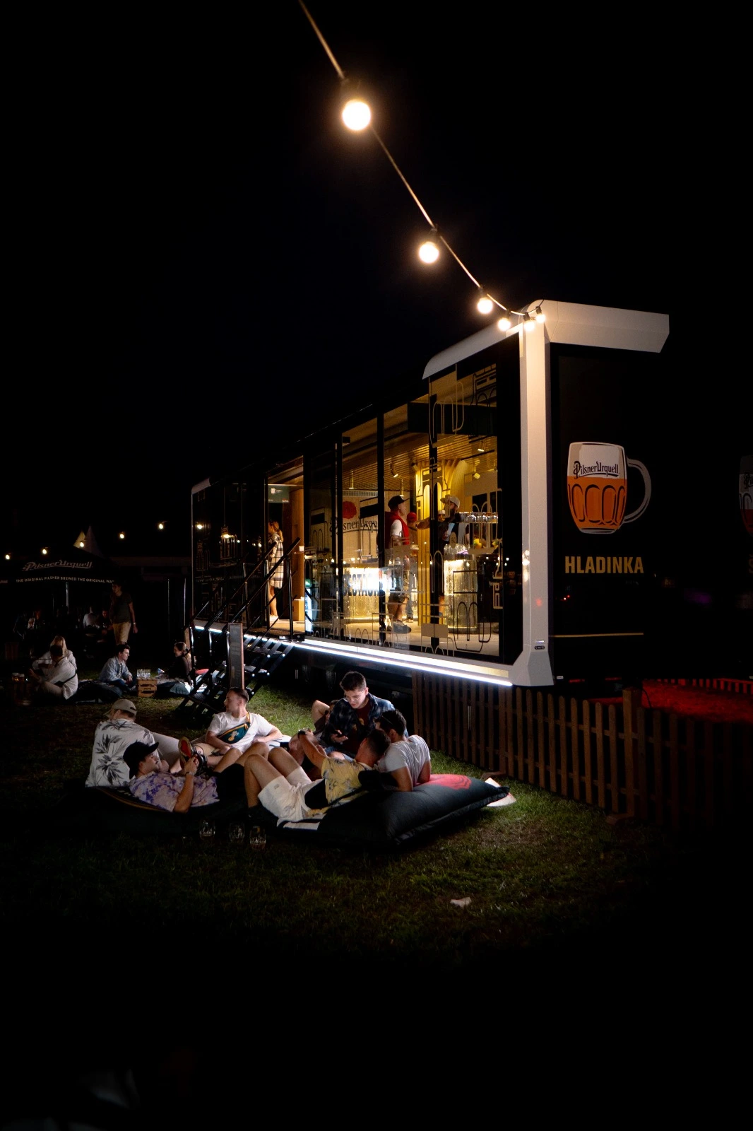 Nighttime outdoor scene of a mobile pop-up bar, with warm lighting illuminating the transparent walls that showcase the interior bar setup.
