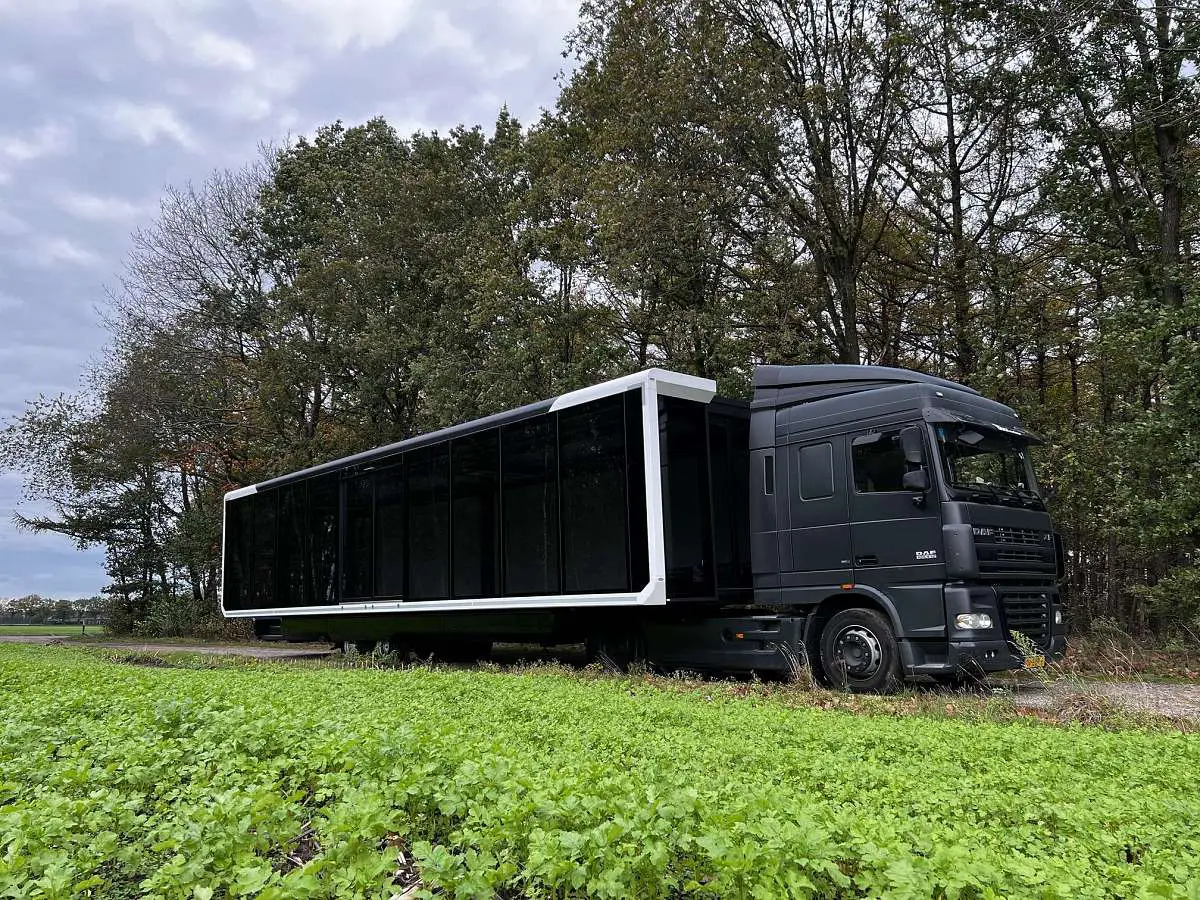 Ein erweiterbarer Baustellen-Büroanhänger, der auf der Wiese neben einem Waldgebiet geparkt ist, mit einer schwarzen Sattelzugkabine an der Vorderseite, was auf eine abgelegene oder ländliche Umgebung für das Baumanagement hindeutet