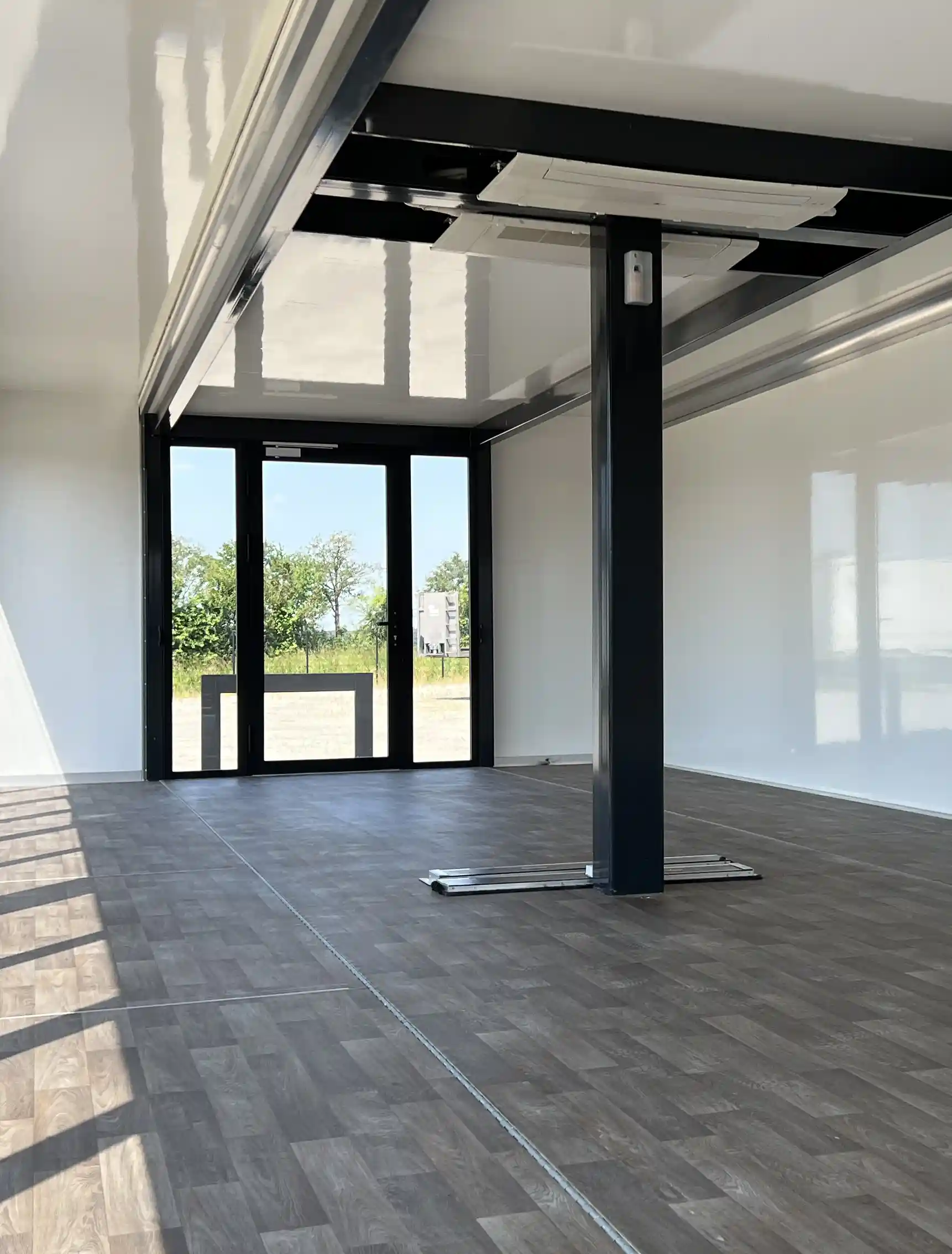 Innenansicht eines erweiterbaren Baustellen-Büroanhängers mit großen Fenstern, die viel Tageslicht bieten, einer zentralen Stützsäule und dunklem holzähnlichem Boden, der auf einen geräumigen und anpassungsfähigen Arbeitsbereich hinweist.