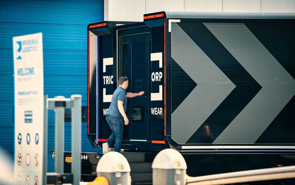A man is opening the door to a black mobile pop-up store with bold "TRICORP WORKWEAR" branding on the side panels. The store is located in an industrial area with a blue building in the background.