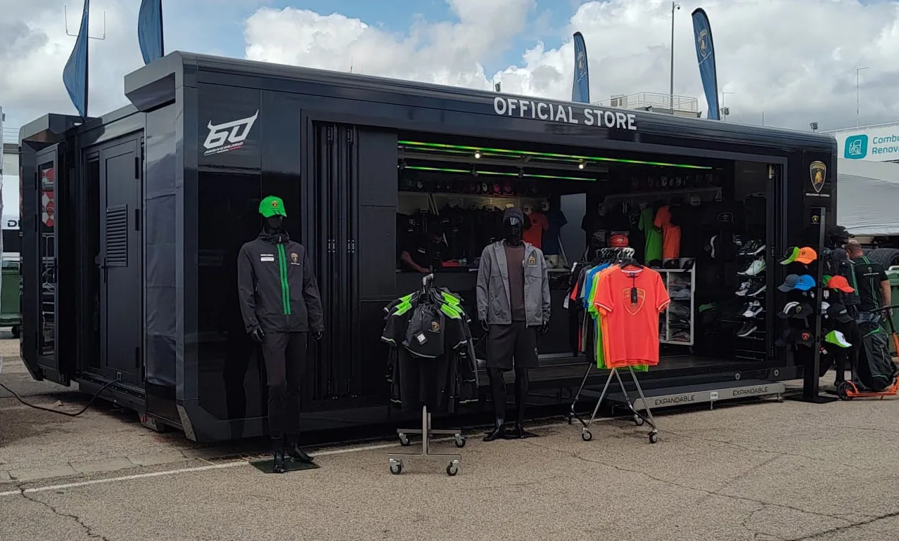 A black mobile pop-up store trailer with large glass windows and "TRICORP WORKWEAR" branding on its side. A man in a hi-vis vest and workwear walks by, indicating active use. The trailer is parked in an industrial area by a building marked "B1".