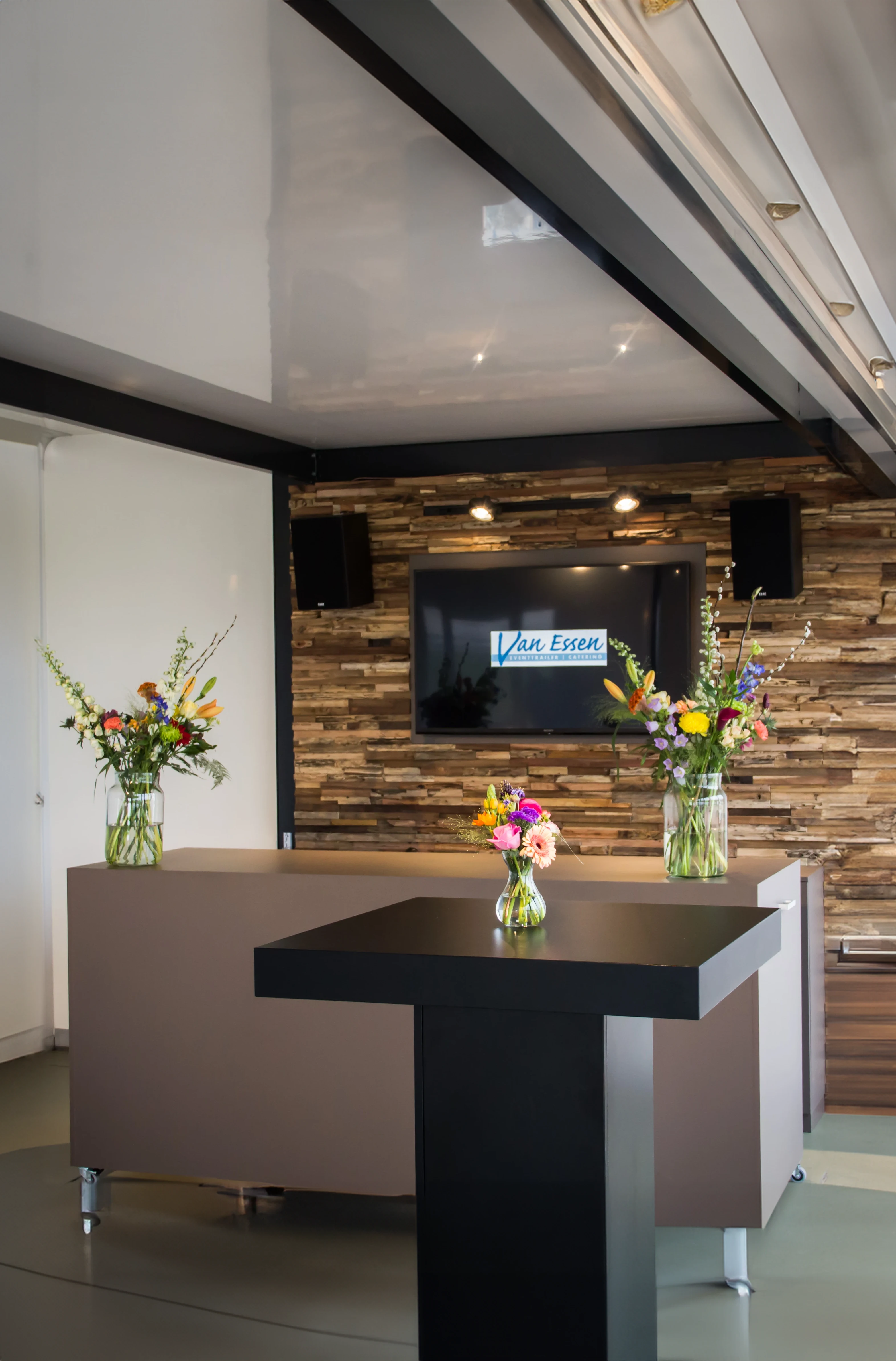The sophisticated interior of a mobile food trailer featuring a reception desk with vibrant flower vases, a wood accent wall, and a mounted TV displaying a welcome message