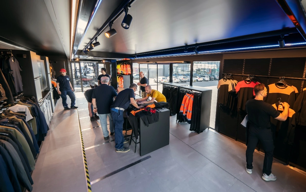 Vue intérieure de la cabine d'essayage mobile Expandable de Tricorp, où les clients et le personnel s'occupent de la sélection et de l'ajustement des vêtements de travail. L'espace bien organisé comprend des étagères de vêtements à haute visibilité et de vêtements de travail décontractés sous un éclairage vif et moderne.