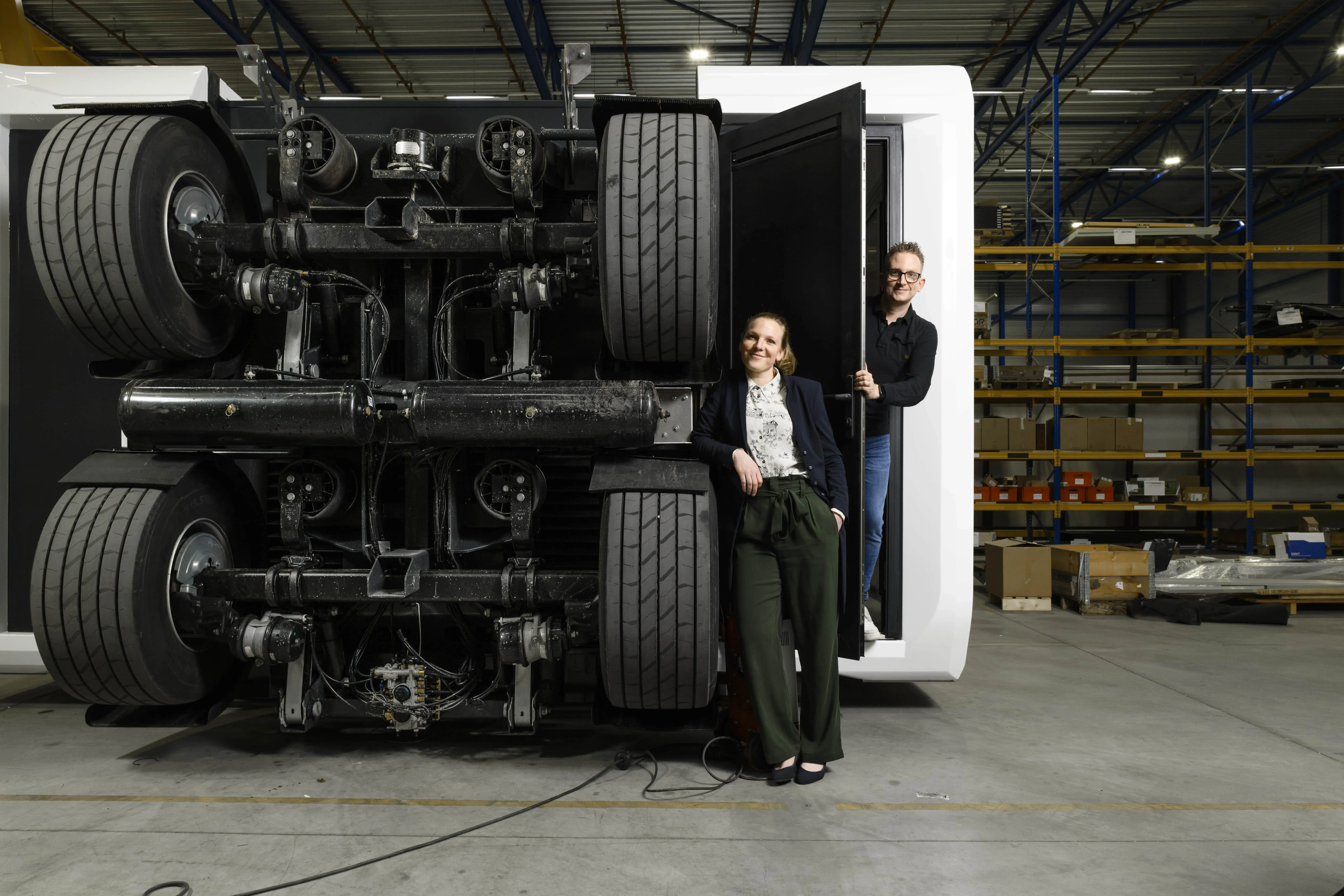 The image shows two individuals standing next to an overturned, large vehicle chassis, presumably for maintenance or educational purposes. The chassis has multiple axles and wheels, indicating it's from a heavy vehicle like a truck or a bus. The setting is an industrial or warehouse environment, with shelving units in the background containing various boxes, suggesting this might be a storage or a workshop area. The presence of the people gives a sense of scale to the large vehicle parts.