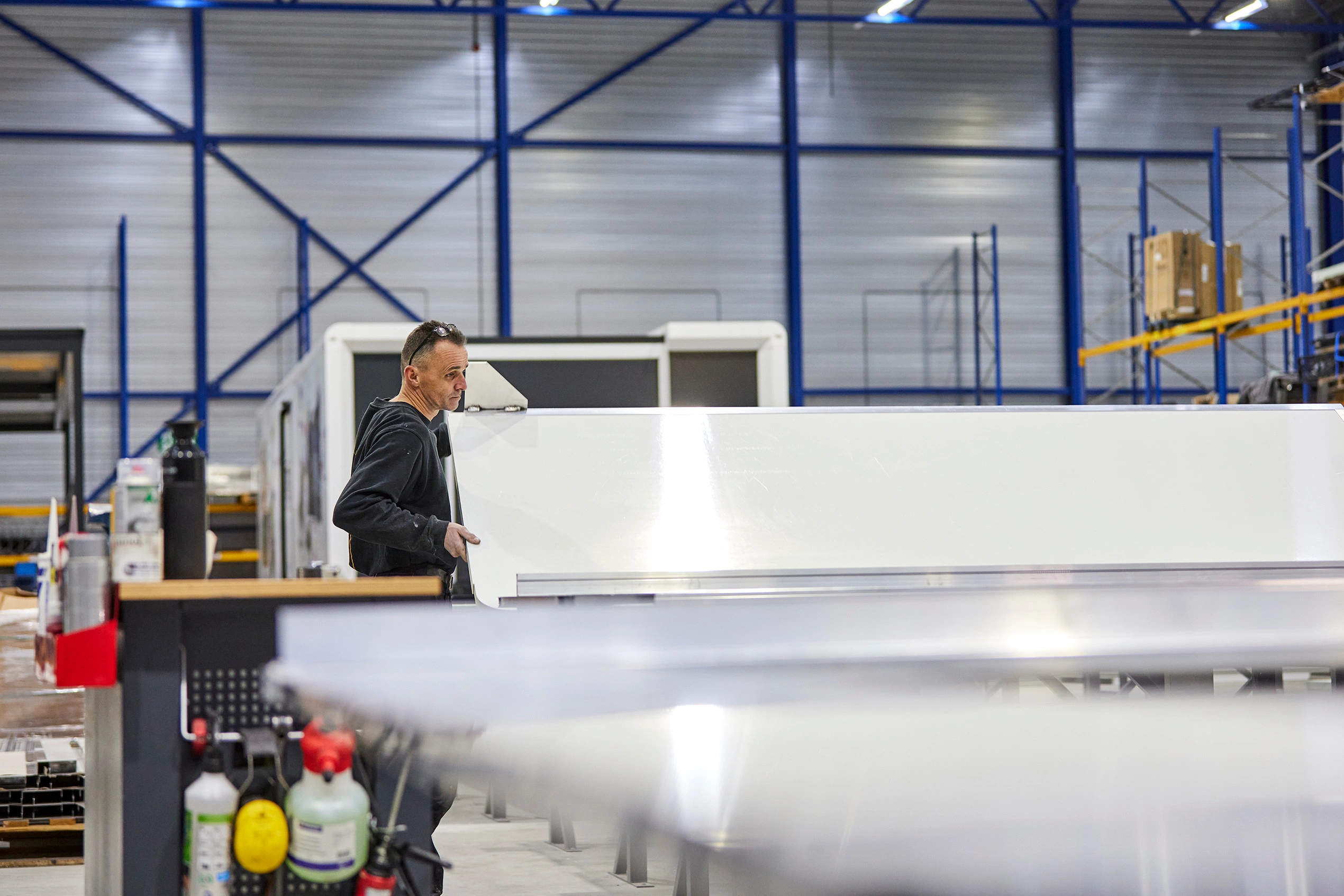 An individual in a black t-shirt and grey trousers is adjusting a component of a large mechanical structure with blue and black parts. The setting suggests an industrial environment, like a workshop for Expandable trailers.