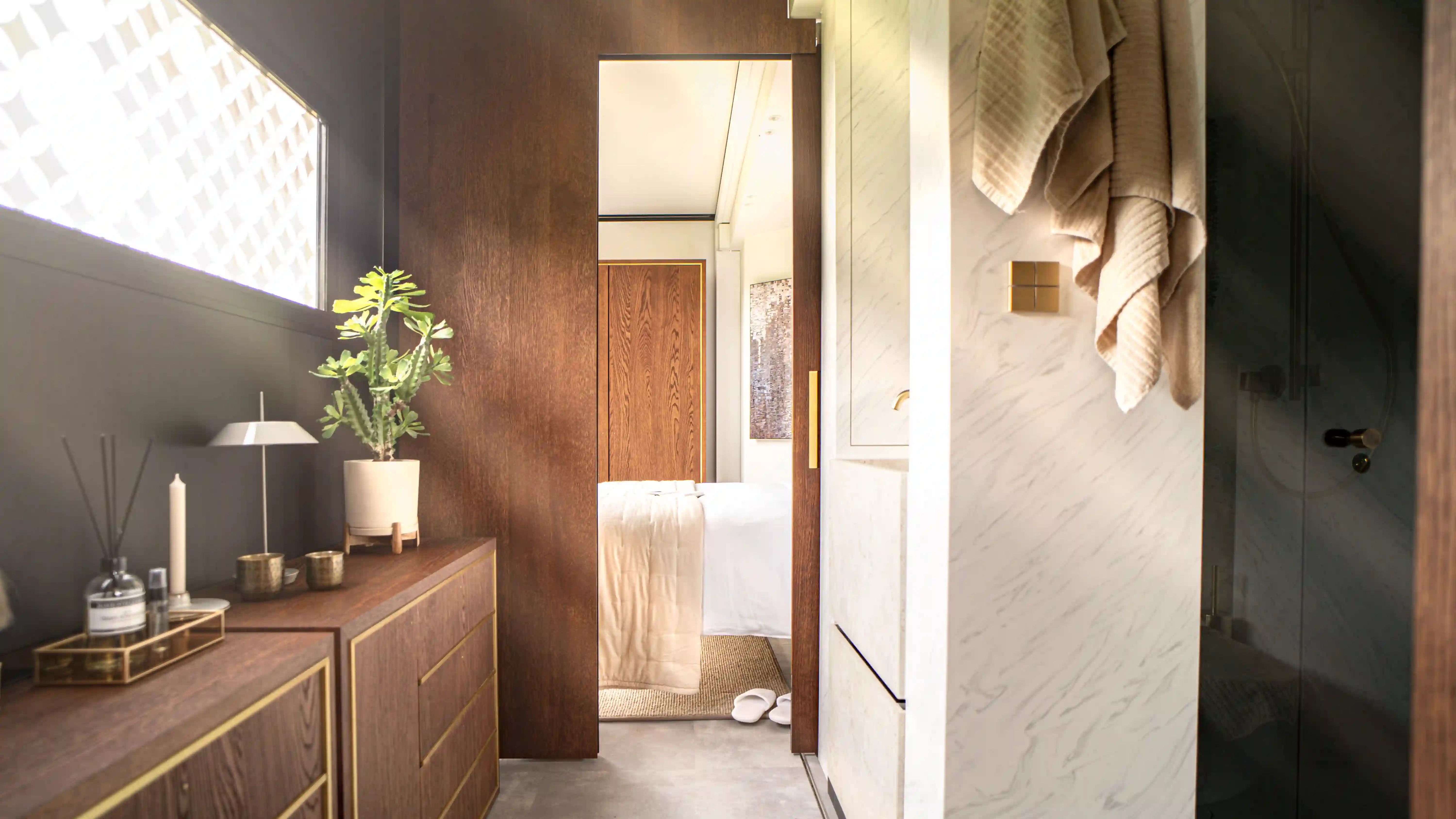 An elegantly designed bedroom in an expandable mansion, featuring a cozy bed with a light comforter, wooden furniture with gold accents, and a plant for a touch of greenery.