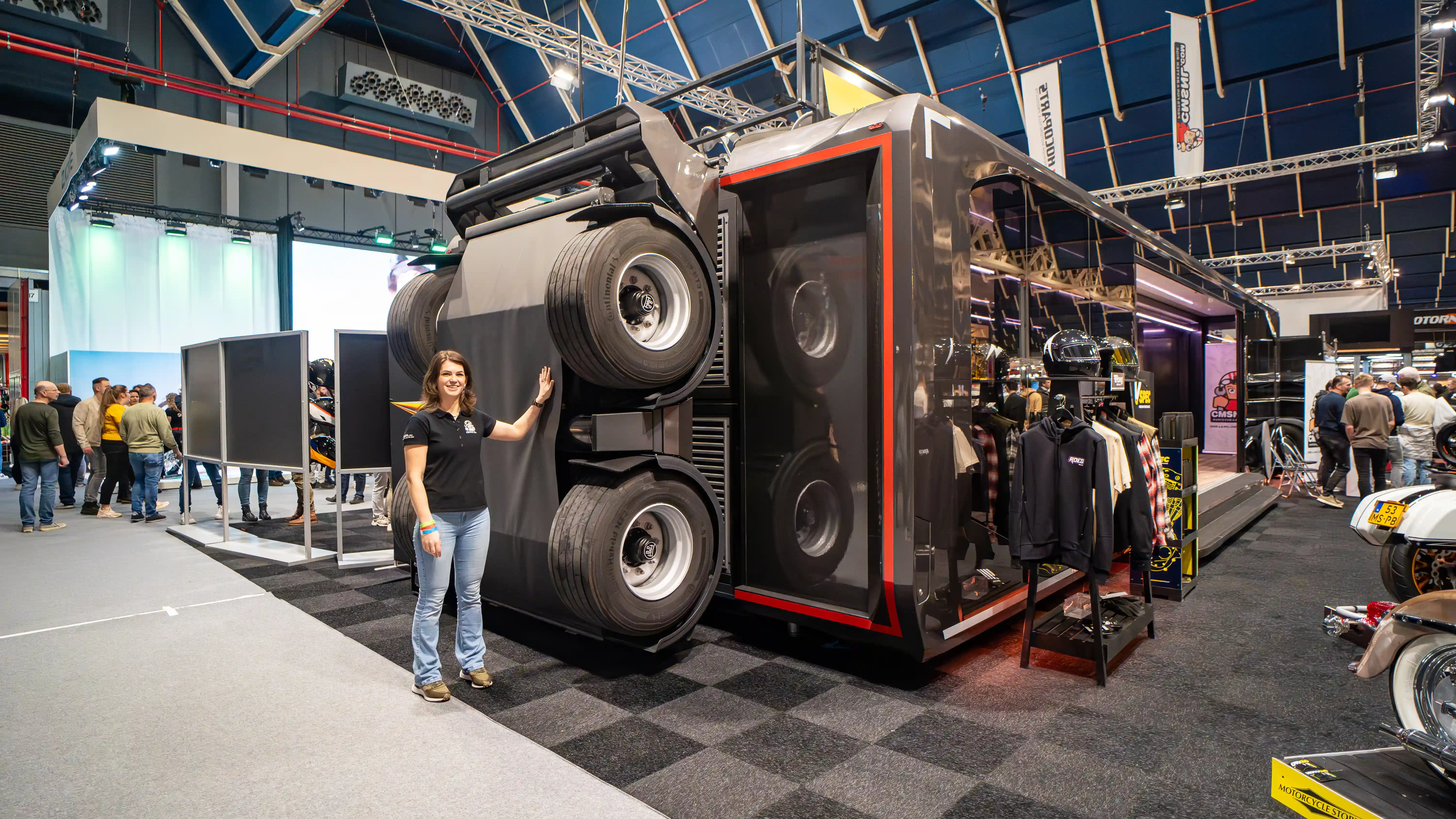 A woman stands beside the lowered Expandable Touchdown trailer at a trade show, showcasing its open side and dual-wheel design, with branded merchandise displayed inside.