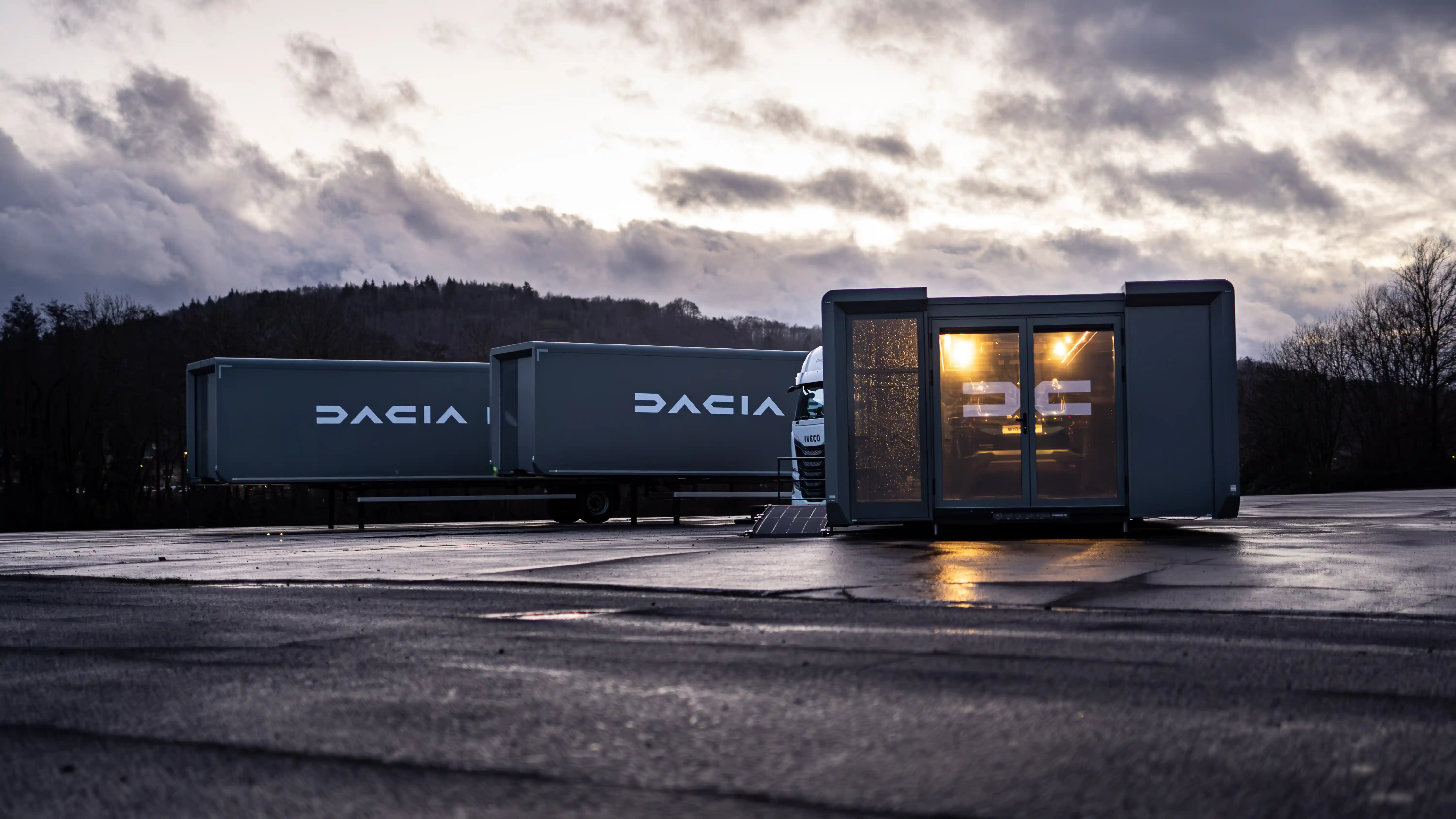 Dawn or dusk setting with a mobile car showroom trailer attached to a black truck parked by a serene lake, the golden hour sunlight reflecting off the vehicle inside the transparent structure.