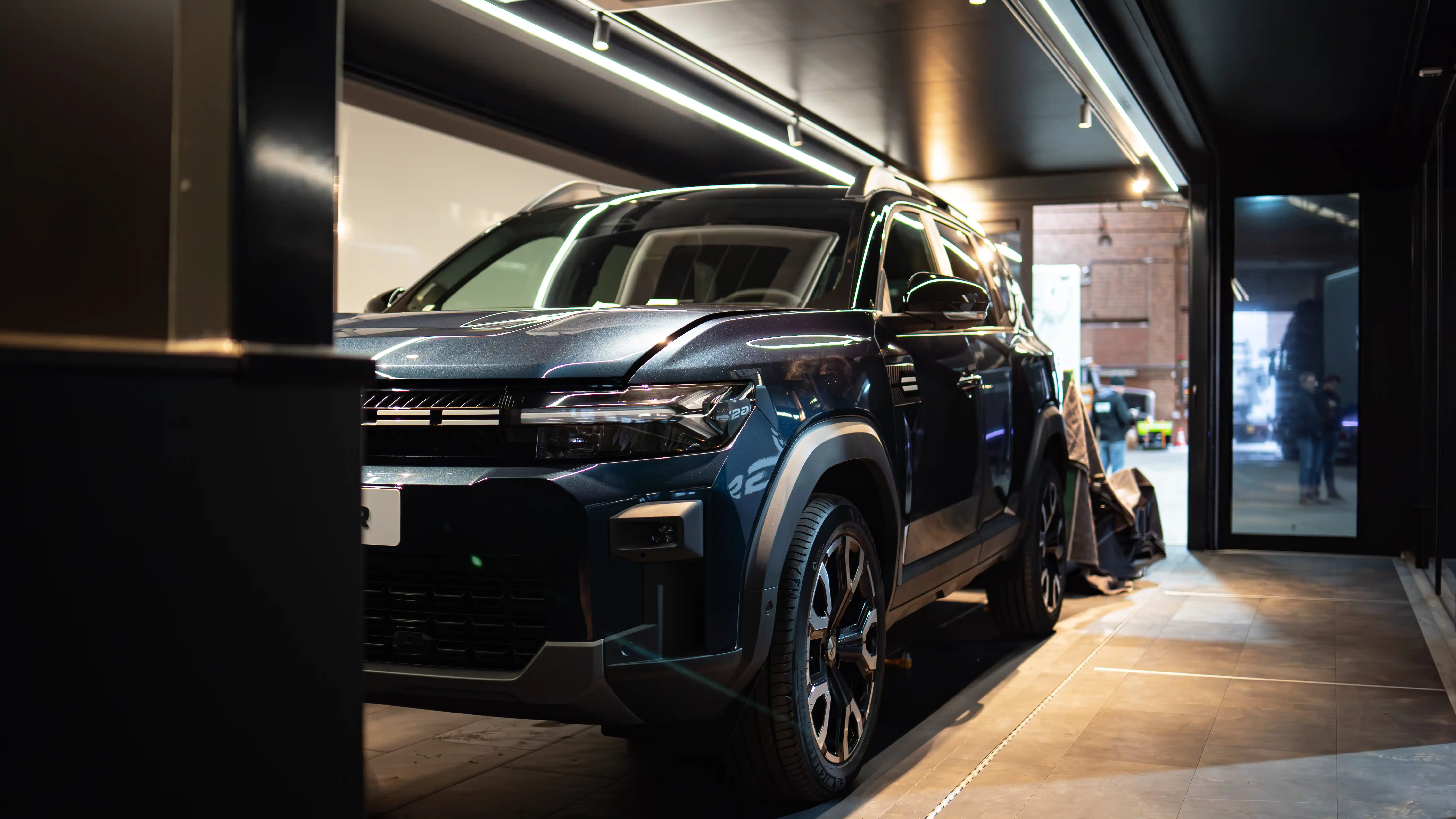 Blick in die Dämmerung auf einen beleuchteten Autohaus an einem Seeufer, in dem ein Luxusauto in einem eleganten, ausziehbaren Anhänger mit Glaswänden ausgestellt ist, der von hohen Pinien flankiert wird.