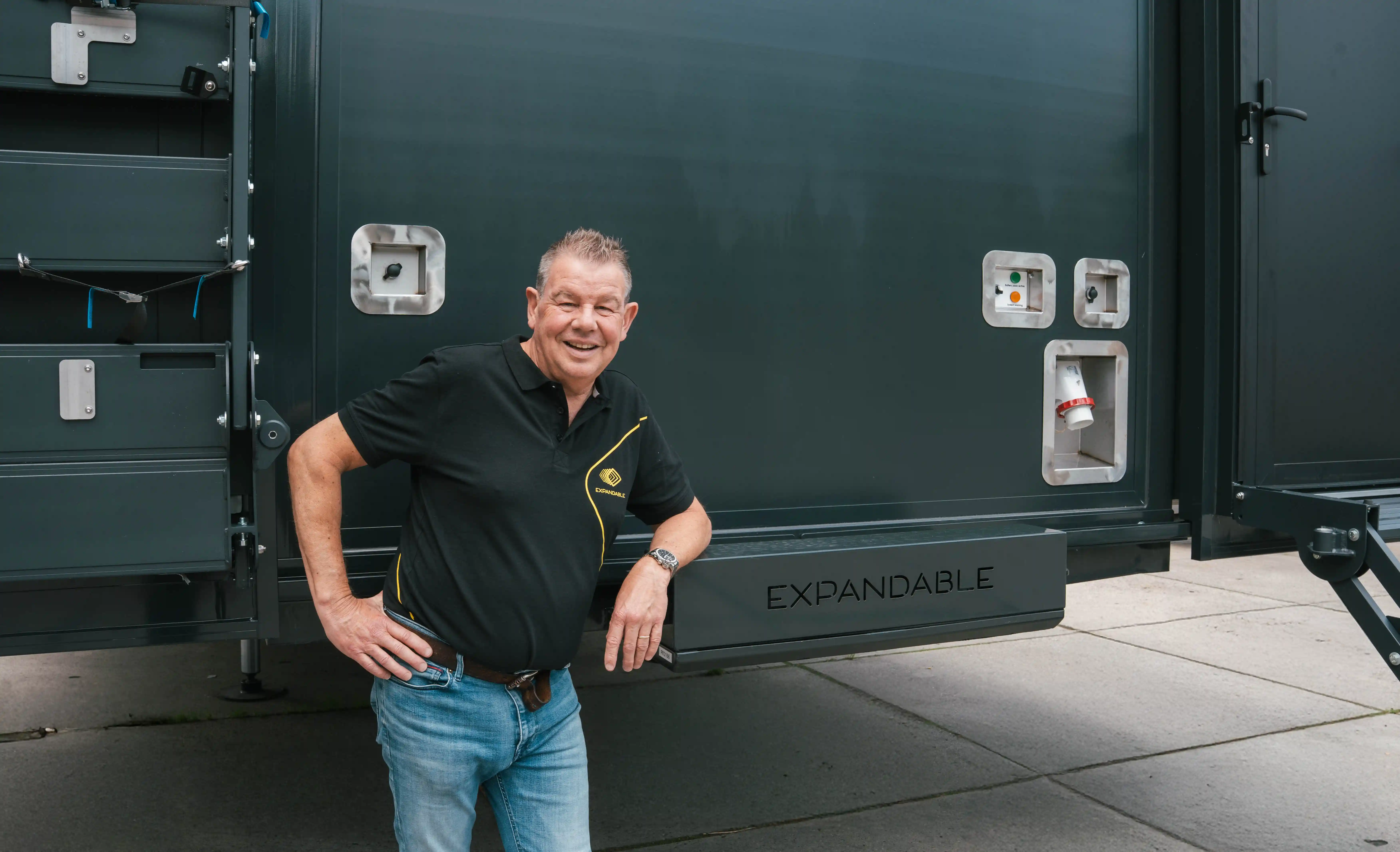 Inside a mobile command trailer with a richly patterned wall and ceiling, technical equipment, and two operator chairs in front of a bank of monitors displaying color bars
