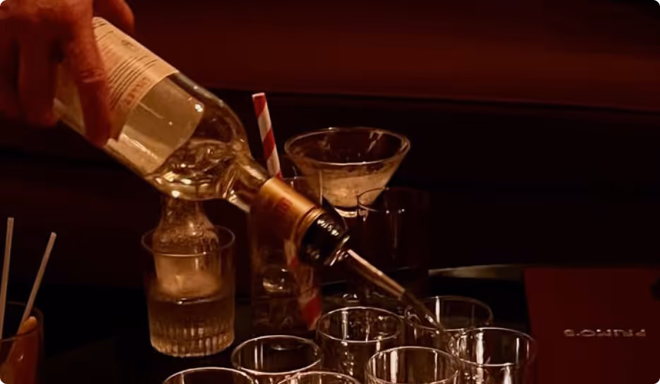 Hand pouring clear liquor from a bottle into multiple empty glasses on a table in a dimly lit setting.