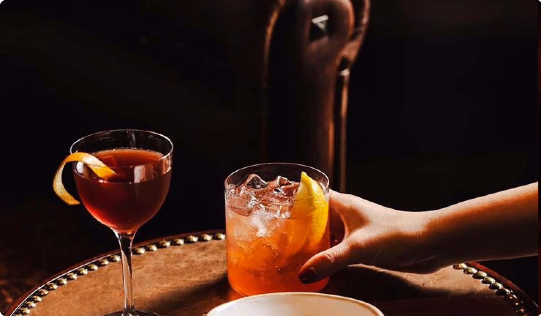 Hand reaching for an iced cocktail with lemon wedge on a round wooden table next to a stemmed glass with an orange twist.