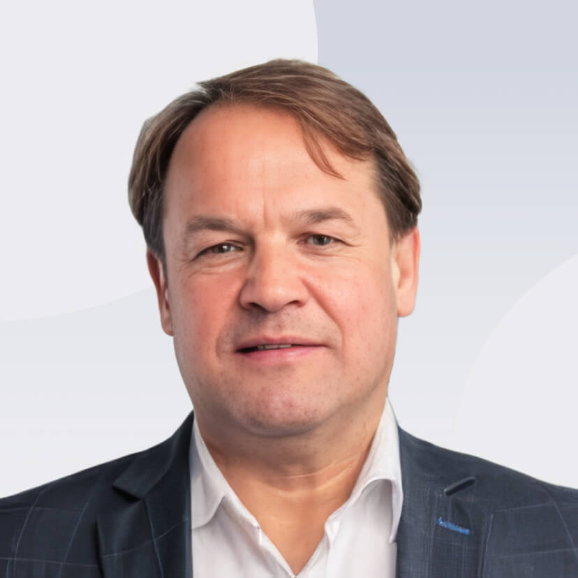 Middle-aged man with light brown hair wearing a dark suit jacket and white shirt against a light background.