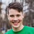 Smiling young man with short brown hair wearing a green shirt outdoors with blurred trees in the background.