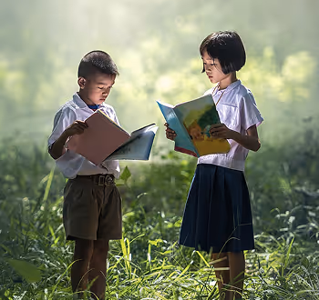Two children in school uniforms standing in tall grass outdoors, reading books in natural light.