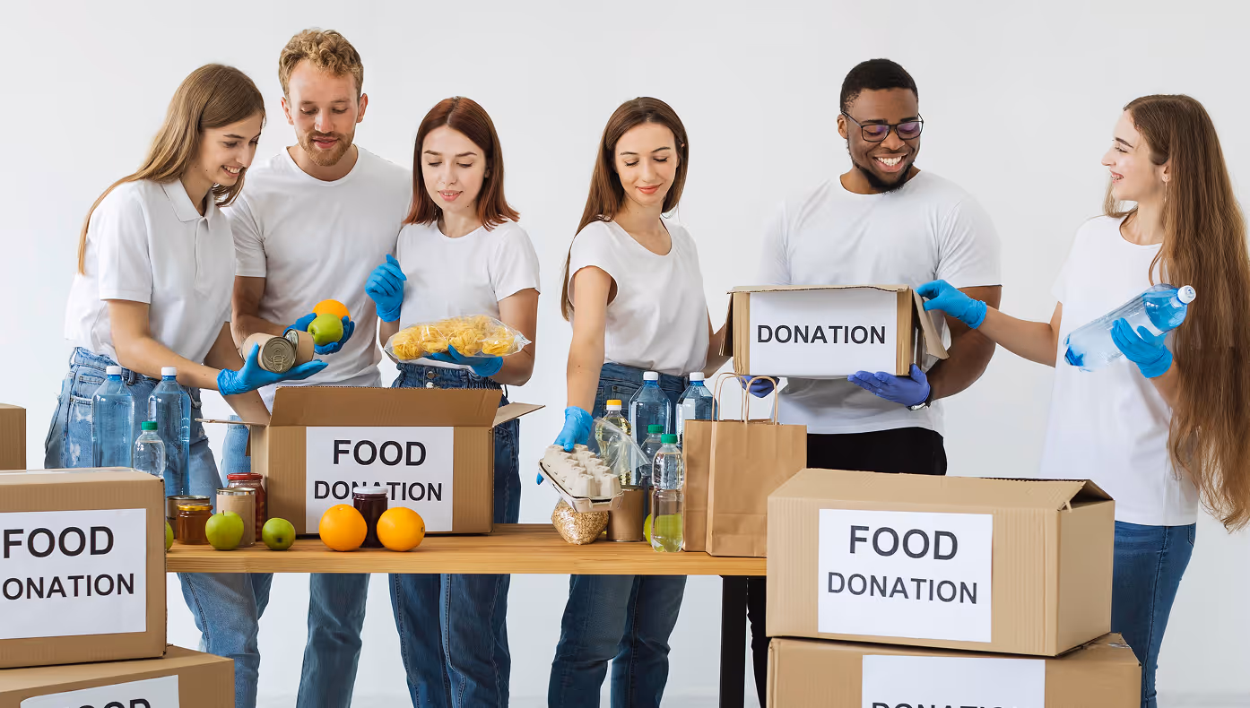 Group of six smiling volunteers packing food and drink donations into labeled cardboard boxes.