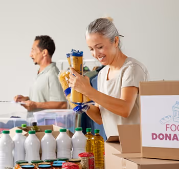 Smiling woman packing food items into a donation box with a man organizing supplies in the background.