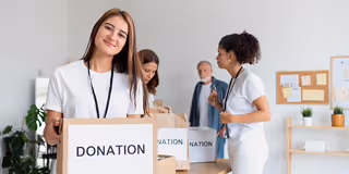 Smiling young woman holding a cardboard box labeled 'DONATION' in a charity setting with volunteers organizing bags in the background.