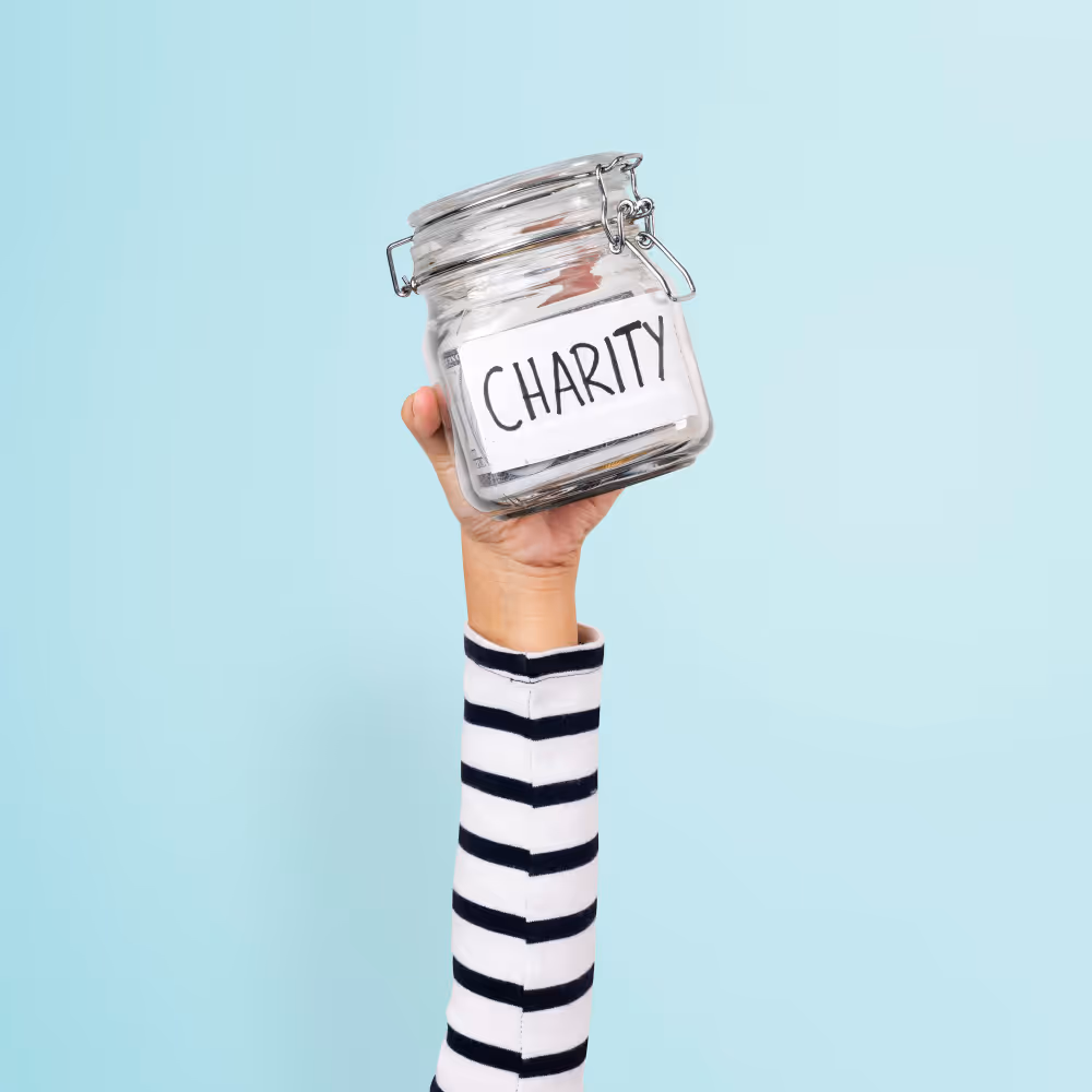 Hand holding a glass jar labeled 'CHARITY' against a light blue background.