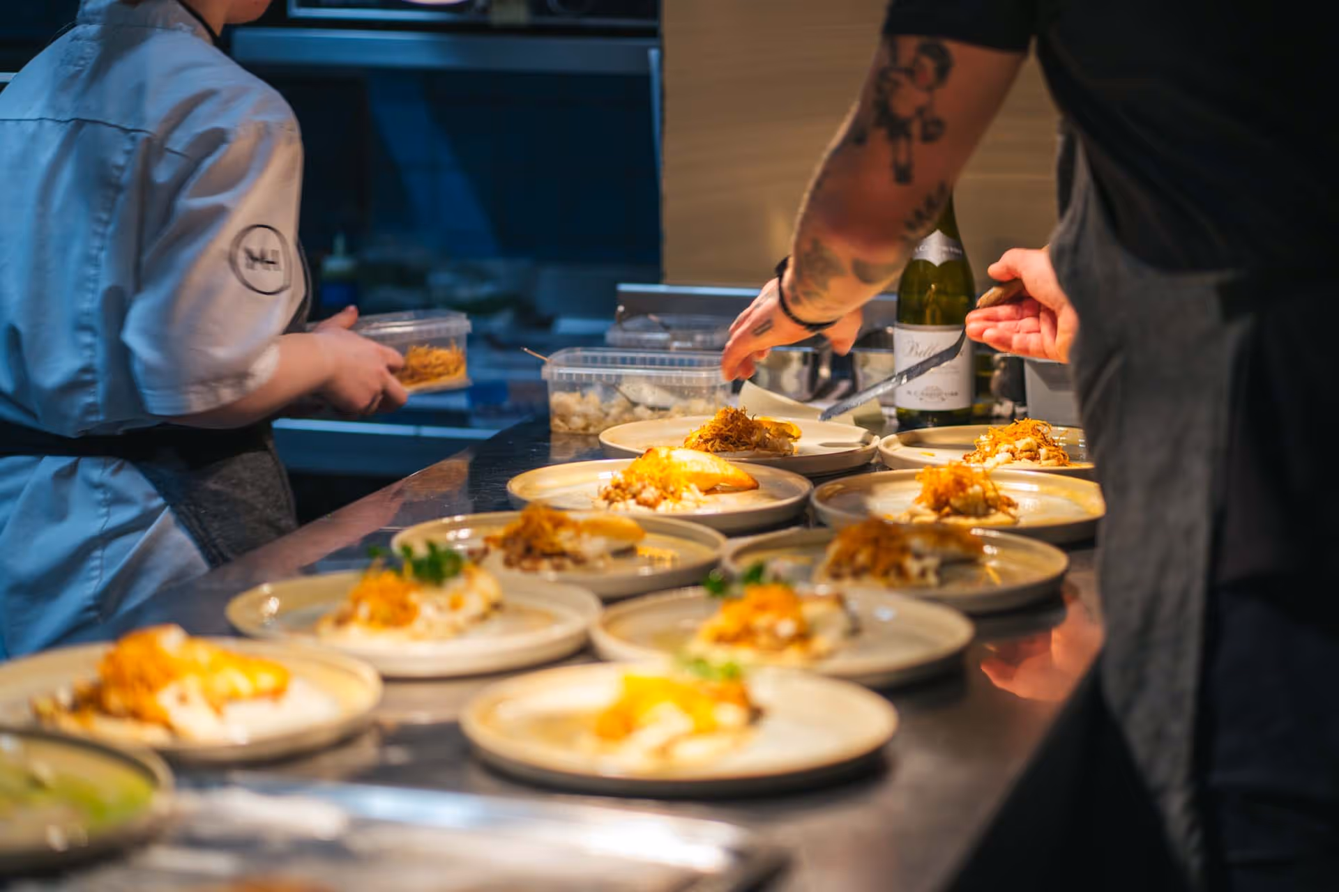 Kokker serverer flere tallerkener med gourmetmat på en rustfri benk i et profesjonelt kjøkken.