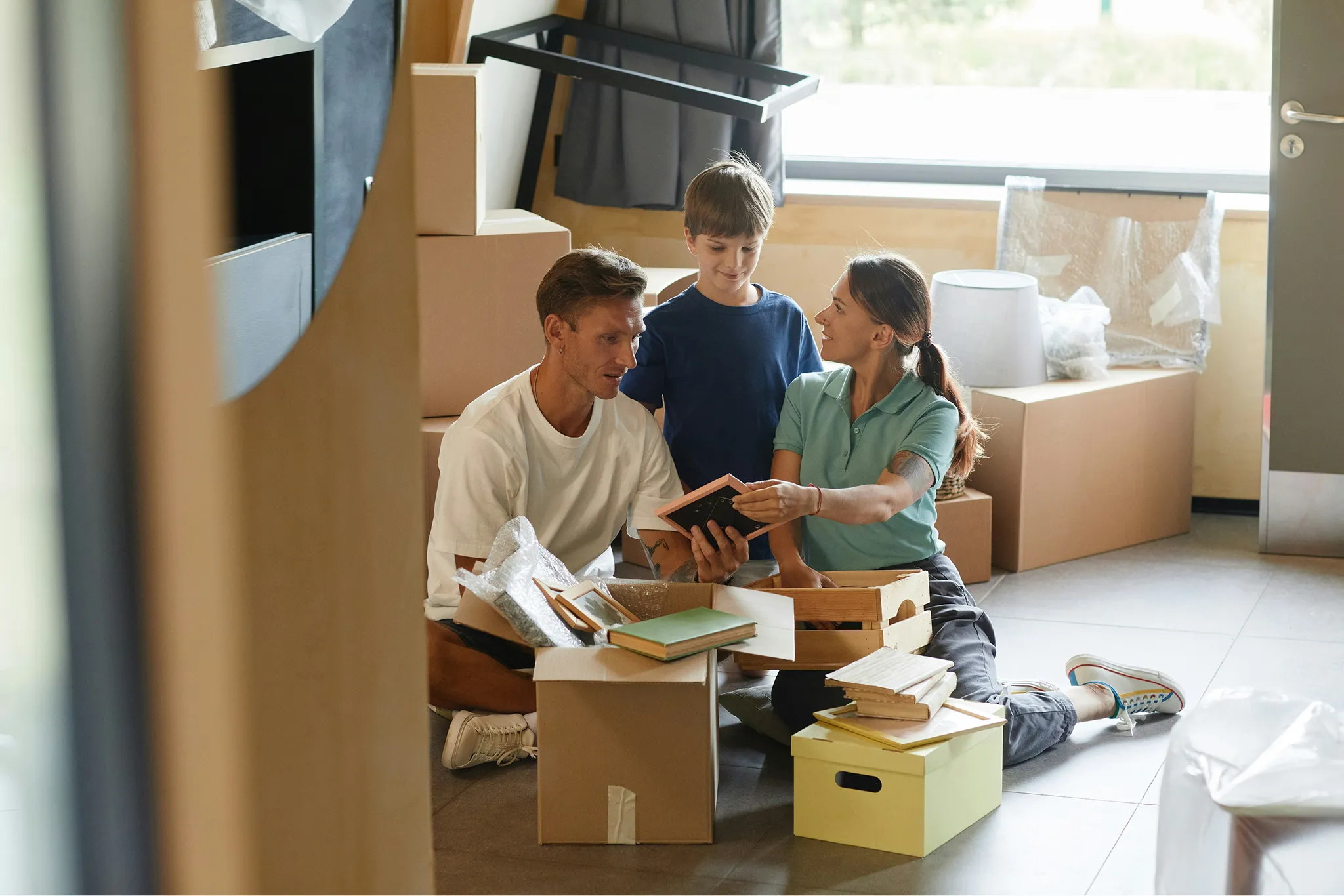 Family unpacking boxes in a new home, sitting on the floor while looking at framed pictures and books.