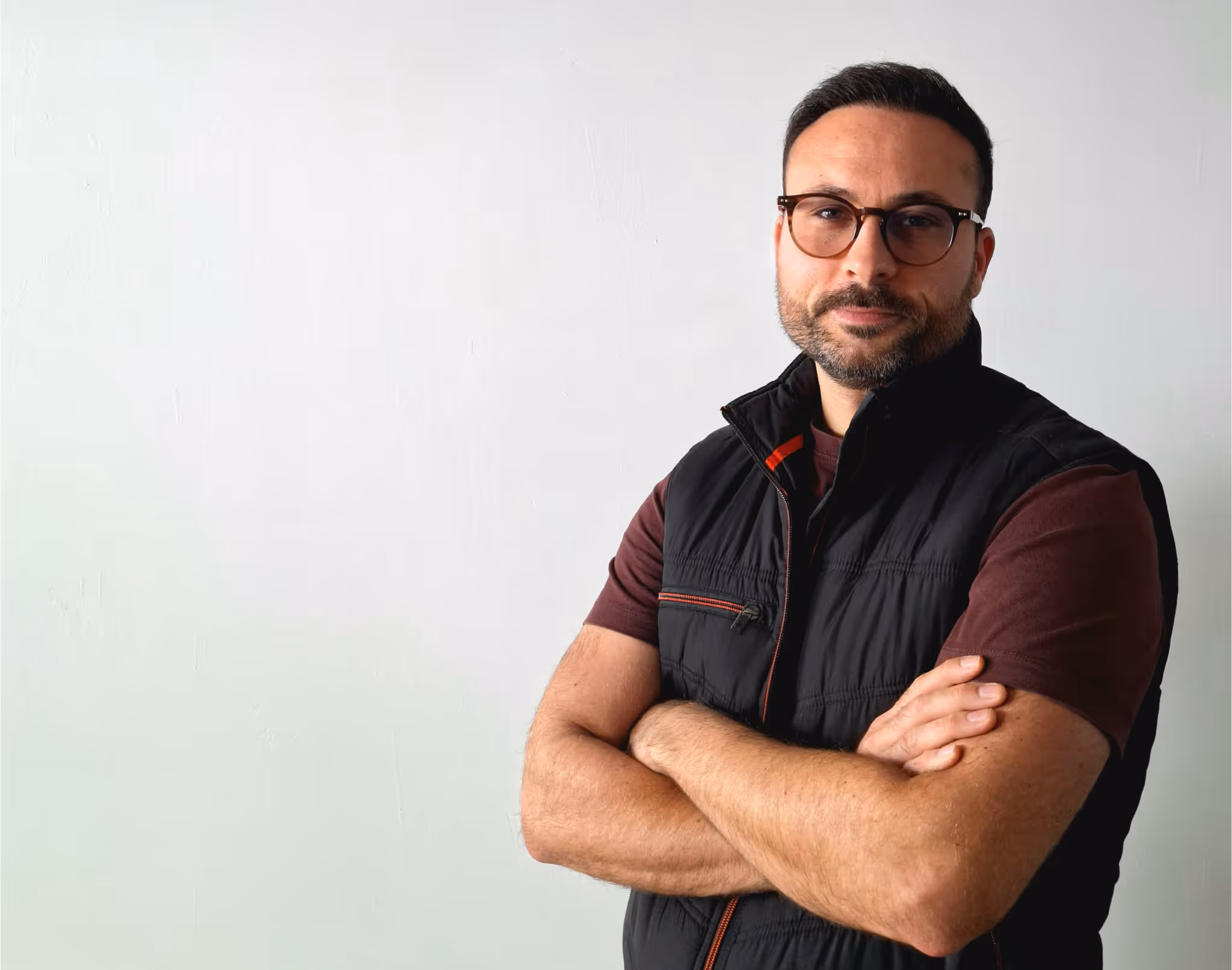 Man with short dark hair and beard wearing glasses, a black vest, and a brown shirt, standing with arms crossed against a plain white wall.