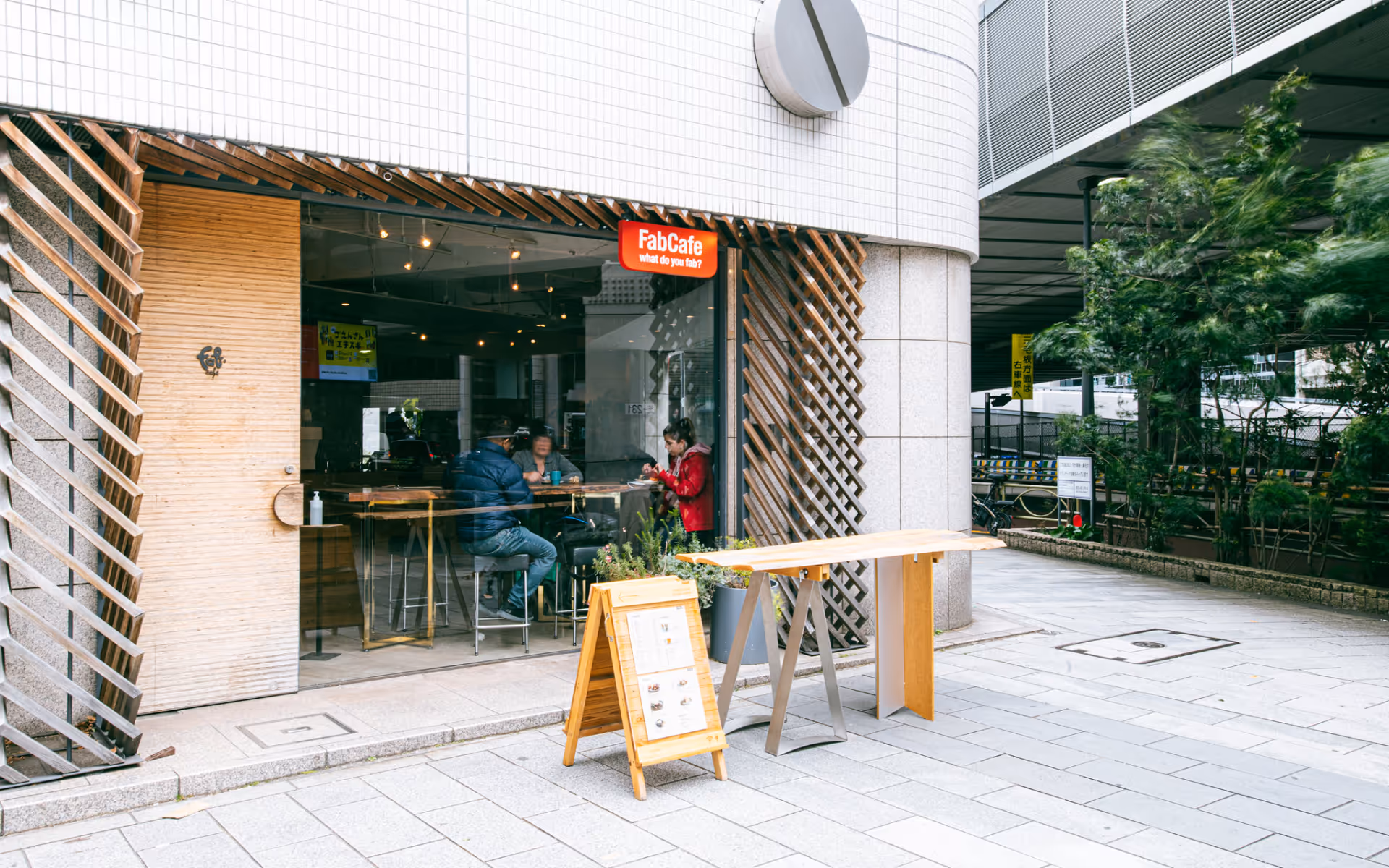 Exterior of FabCafe Tokyo with glass storefront, wooden façade details, and people seated inside.
